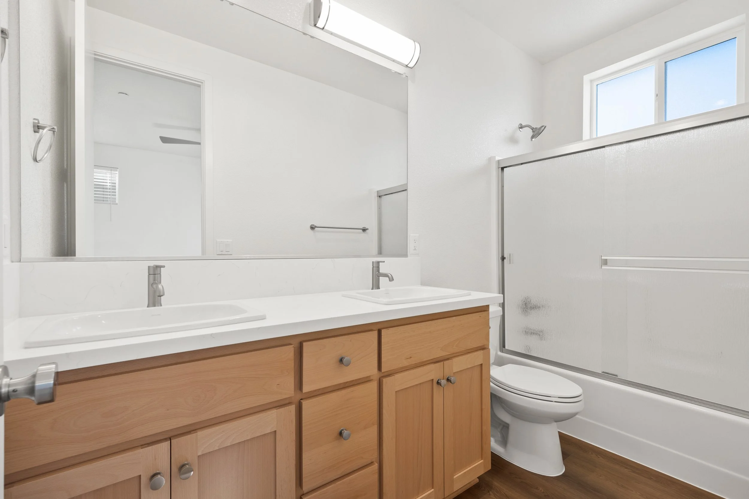 Bathroom with double sink vanity, large mirror, shower with sliding frosted glass doors, toilet next to vanity, window, and wood flooring.