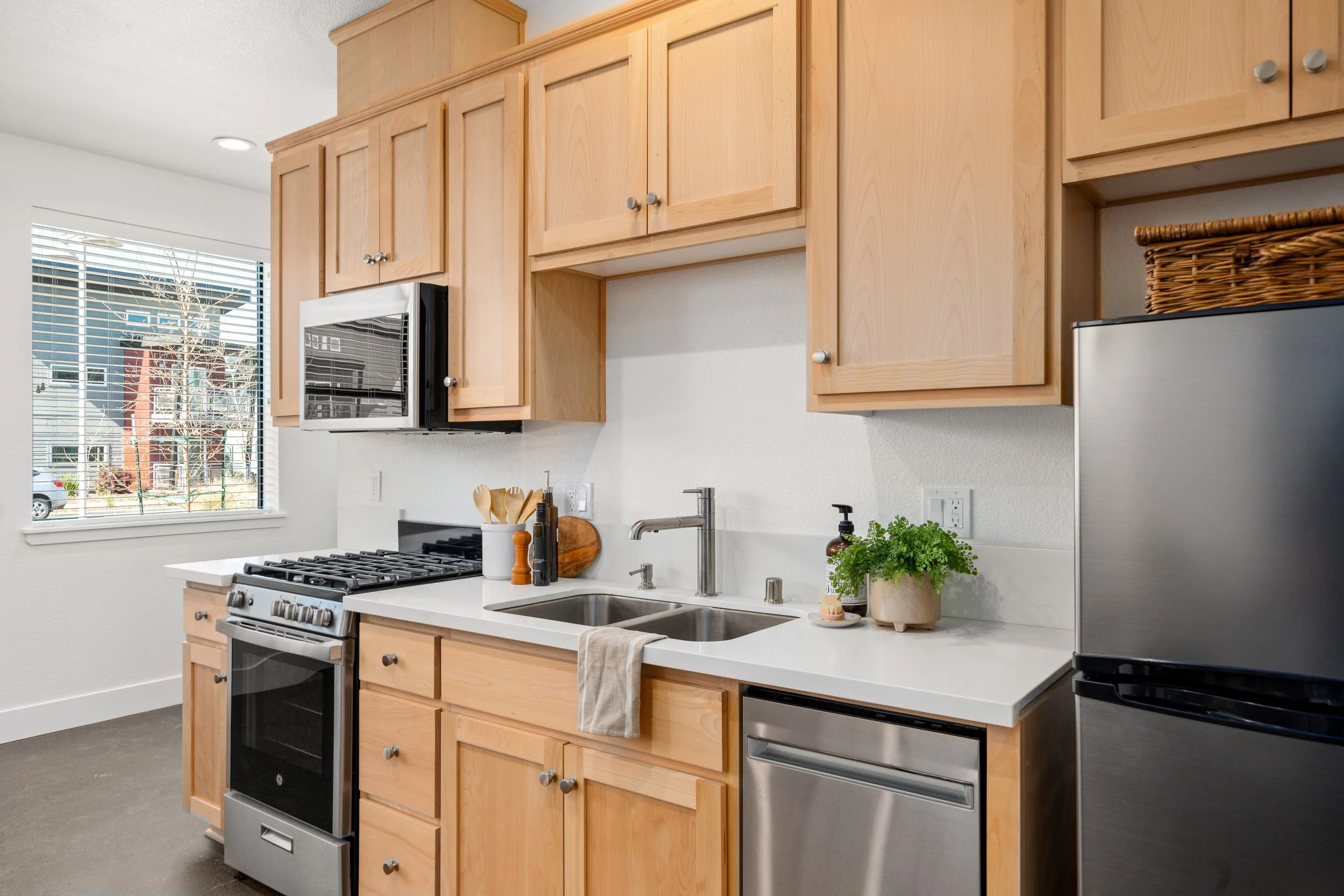 Modern kitchen with wooden cabinets, stainless steel appliances, a window with blinds, and green potted plant on the counter.