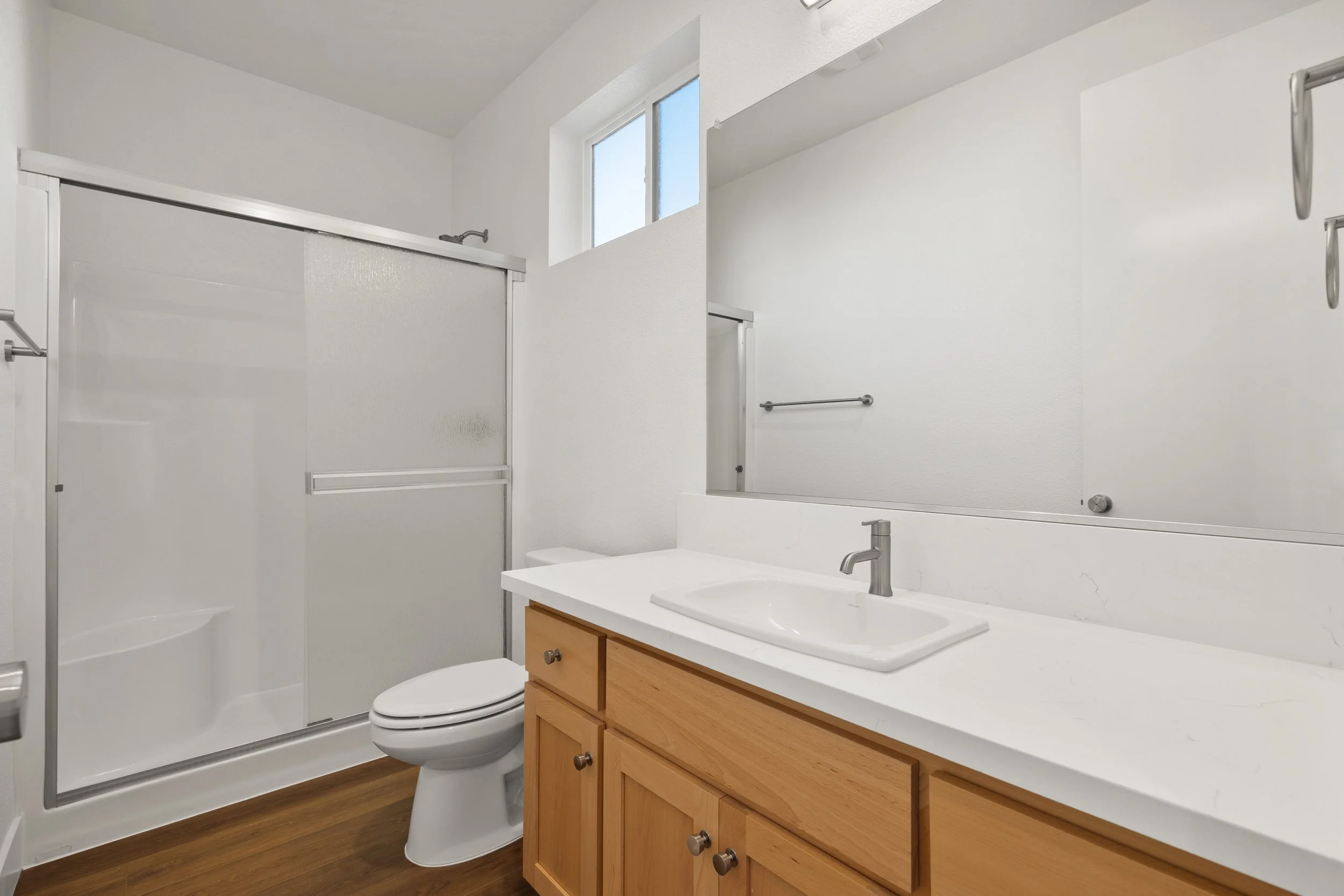 A small bathroom with a shower with frosted glass sliding doors, a white toilet, a wooden vanity with a white countertop and sink, and a large mirror above the sink. The bathroom has white walls, a small high window, and wooden flooring.