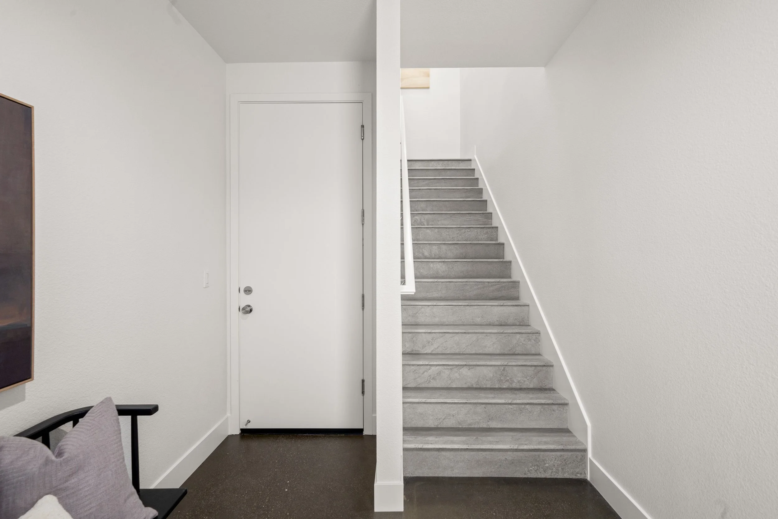 Interior view of a hallway with a white door, a staircase with concrete steps, and a small portion of a black bench with purple cushions.