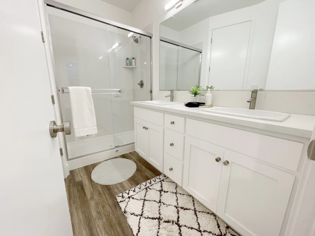 A modern bathroom featuring a glass-enclosed shower with white tile, a white vanity with a large mirror, a potted plant, soap dispenser, and a patterned bathmat on wood-look flooring.