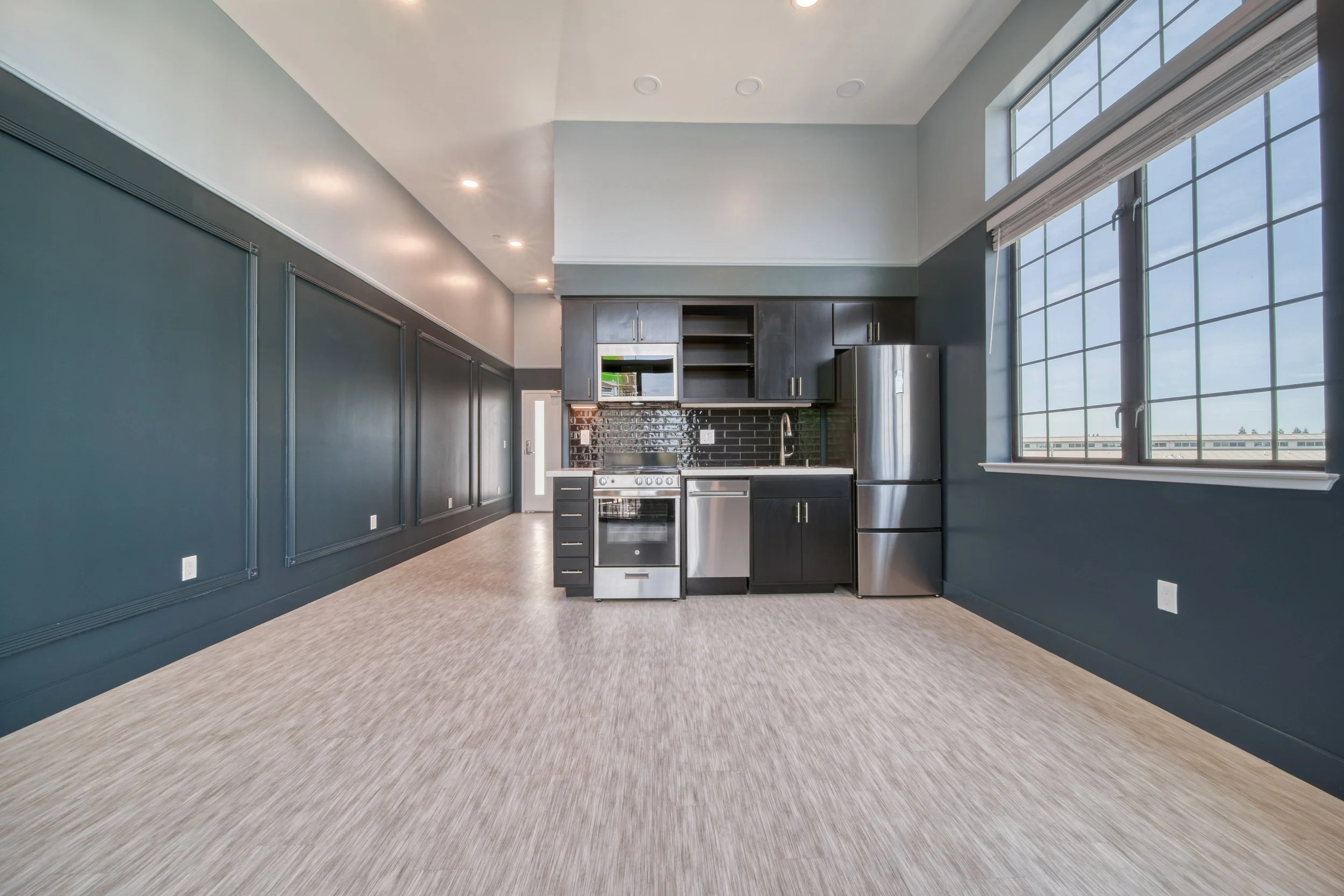 Empty modern kitchen with dark cabinets, stainless steel refrigerator, stove, and dishwasher, large window with blind, beige carpet flooring, and blue paneled walls.