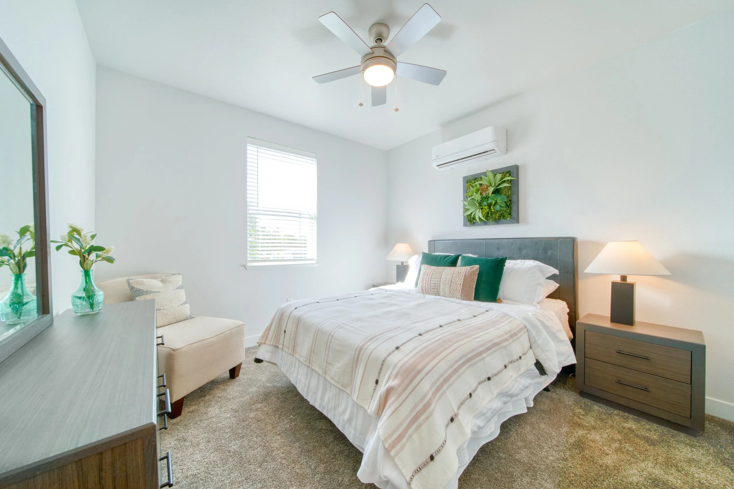 A bright bedroom with a white ceiling fan, window with blinds, air conditioner, and a wall art piece of greenery above the bed. The bed has white and green pillows, a beige and white striped bedspread, and a dark gray headboard. There are two matchin