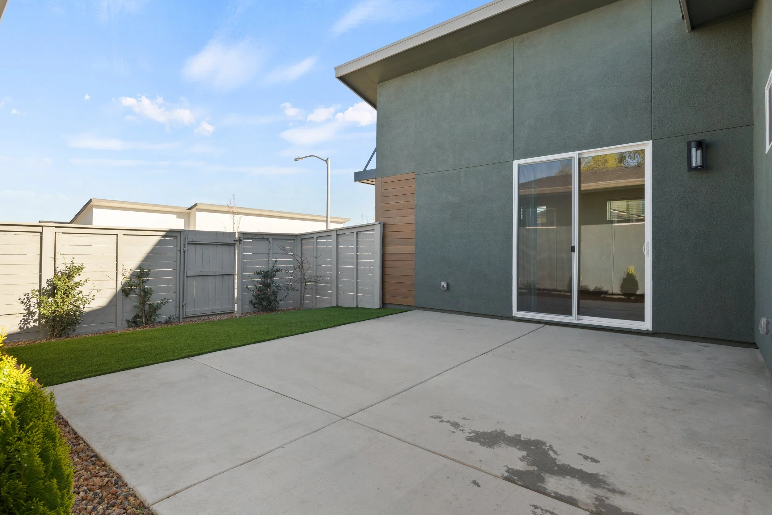 Modern backyard with concrete patio, small green lawn, a white wooden fence, and an exterior house wall with a sliding glass door