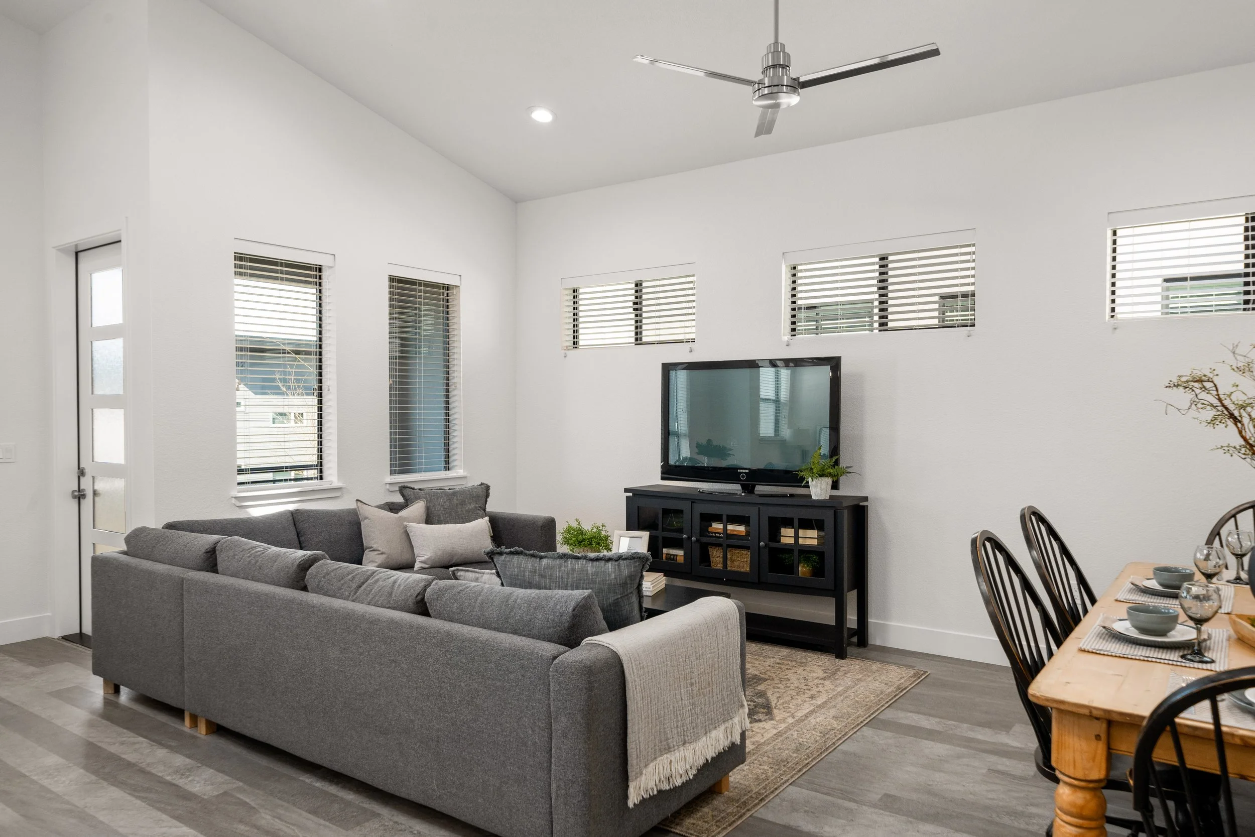 Modern living room with gray sectional sofa, wooden dining table, black TV stand, flat-screen TV, and multiple windows with blinds.