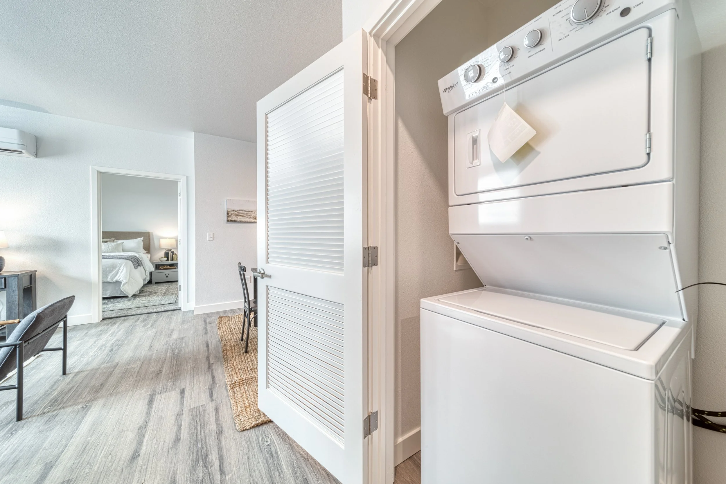A laundry closet with a stacked white washing machine and dryer next to a door with louvered panels, open to a living area with a bedroom visible in the background.