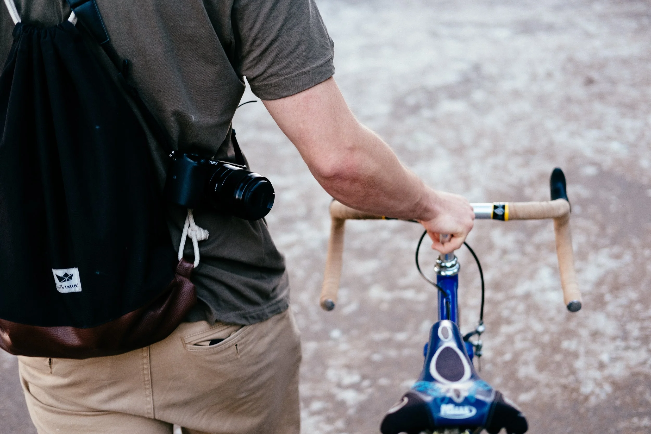 Person holding a small bicycle on a gravel surface, with a camera hanging from their neck.