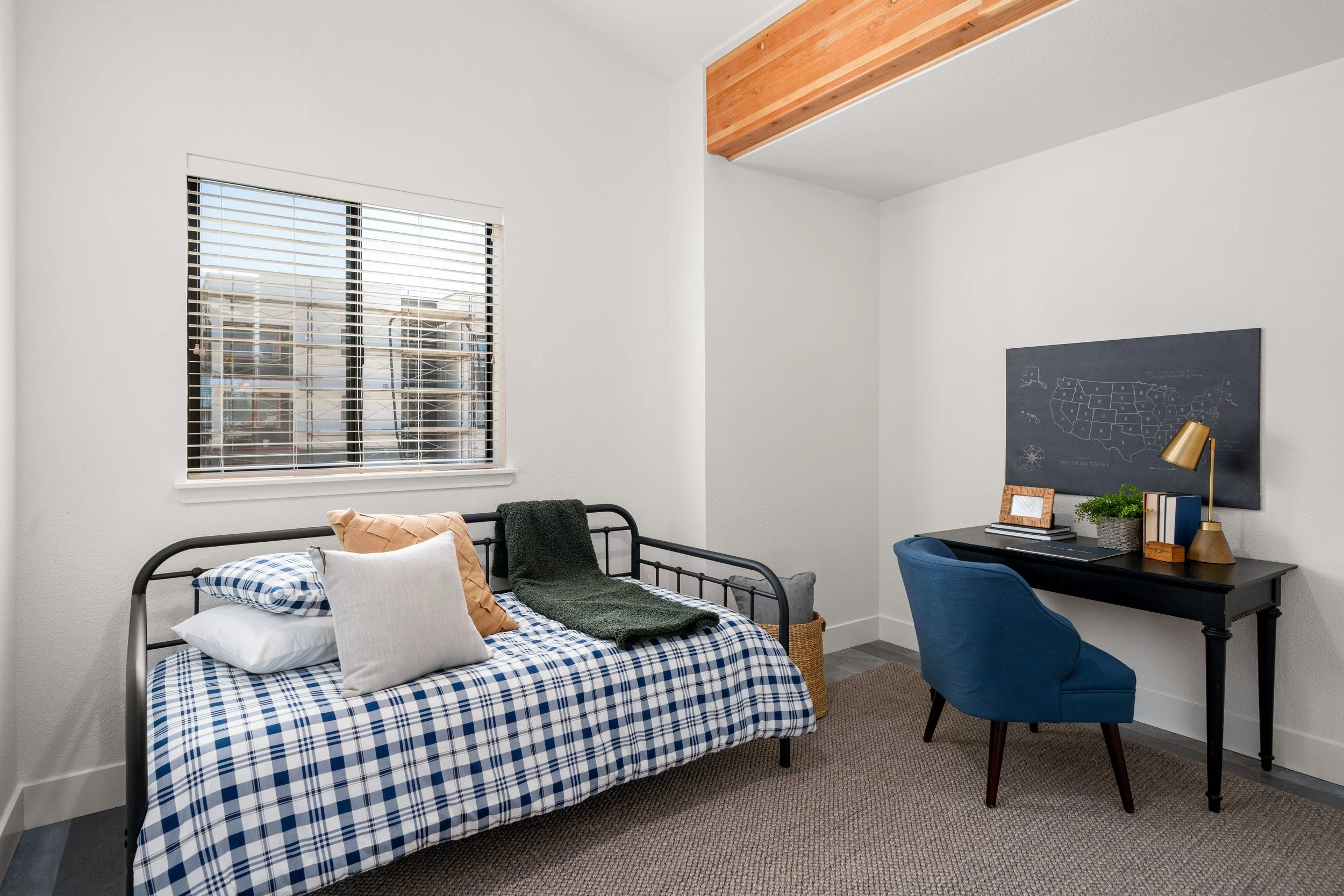 A bedroom with a window, a black metal daybed with plaid bedding, pillows, and a blanket, a black desk with a blue upholstered chair, a table lamp, books, and decorative items, beige carpet, white walls, and wooden ceiling beam.