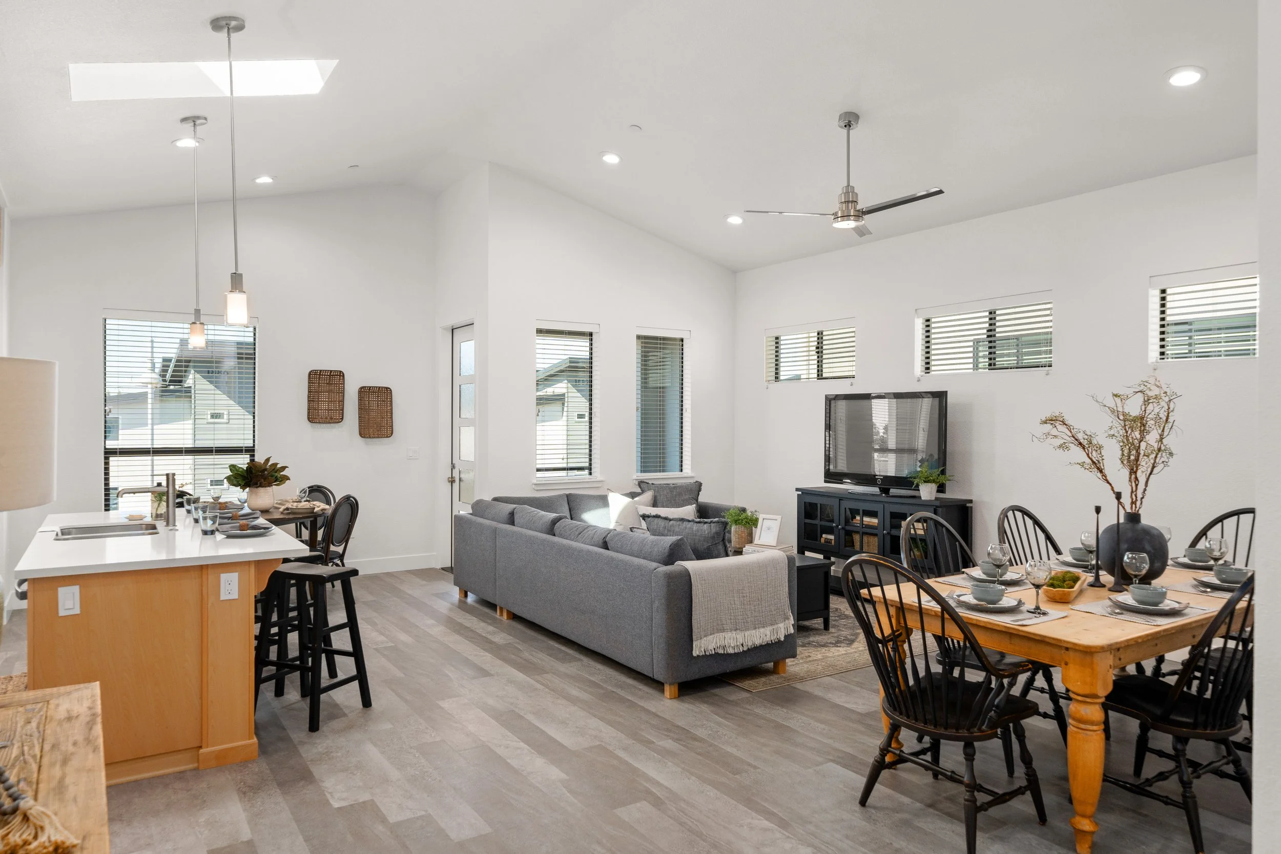 Open-concept living and dining area with white walls, multiple windows, a gray sofa, a wooden dining table with black chairs, a TV on a black stand, and kitchen island with two black stools.