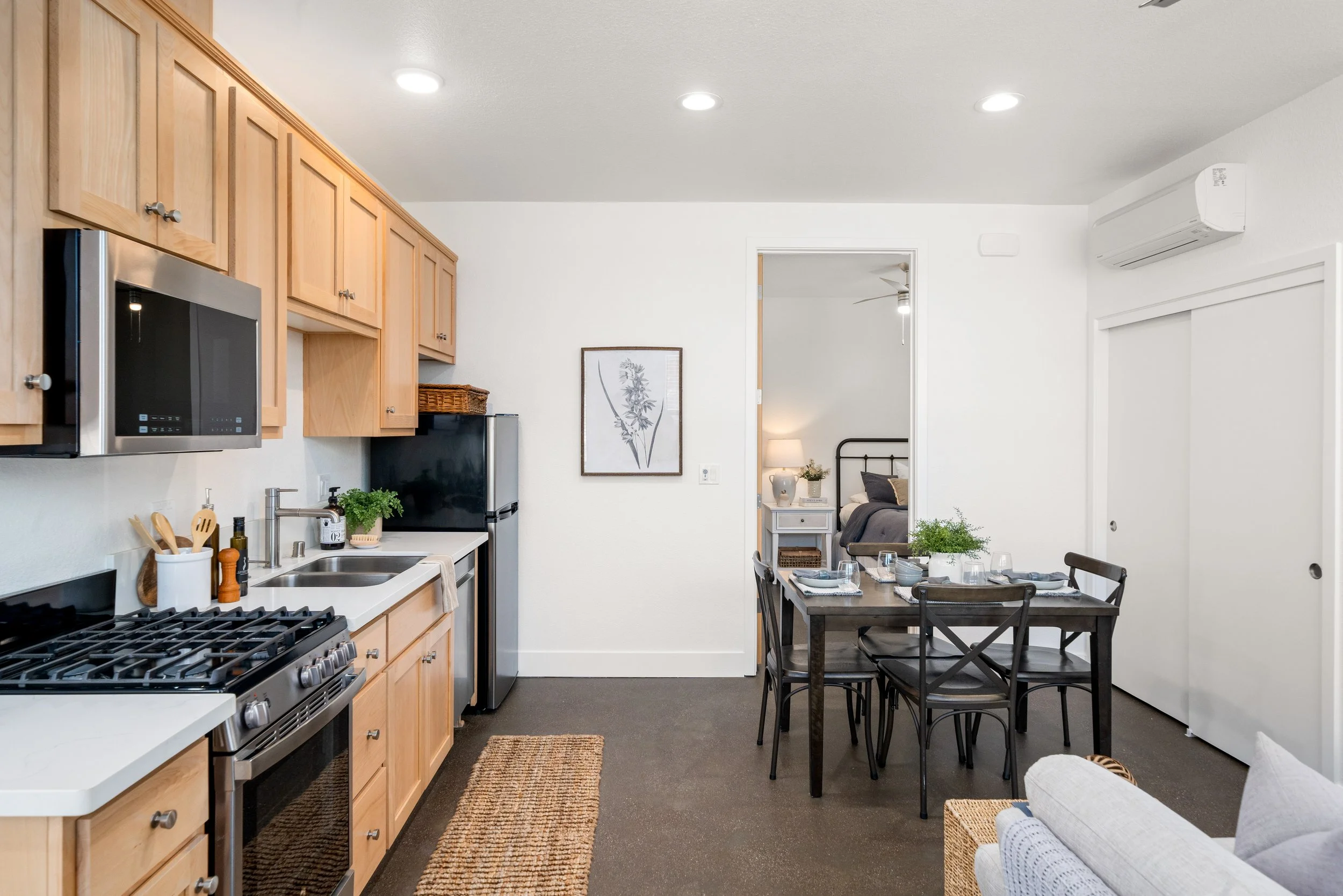 Open-concept living room and kitchen with wooden cabinets, stainless steel appliances, dining table set for four, and a bedroom visible through a doorway.