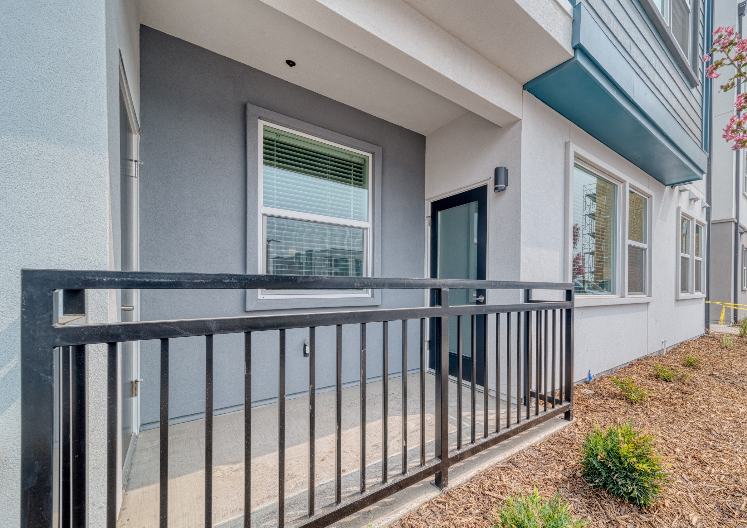 Front porch of a modern apartment building with a black metal railing, white trim around window and door, gray exterior wall, and greenery in the landscaped area.