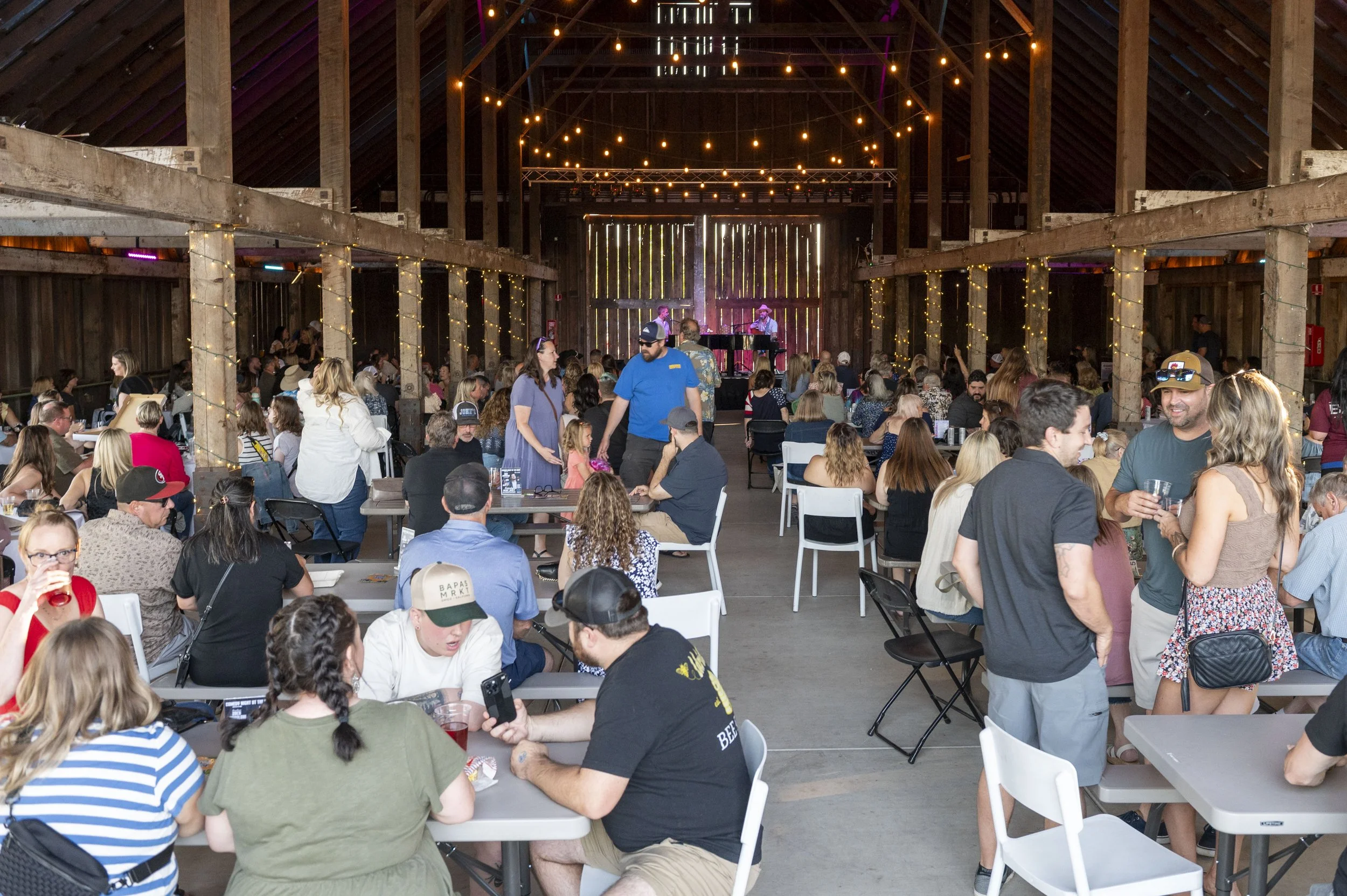 A lively indoor event with many people seated at tables, some standing and chatting, with a stage at the back where musicians are performing. The venue has string lights hanging from the ceiling and wooden walls, giving a warm, rustic atmosphere.