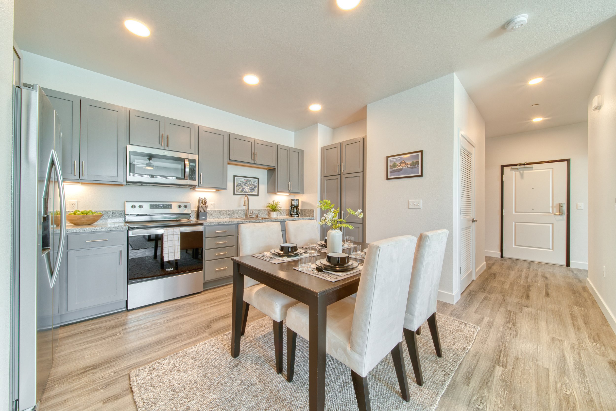 Open-concept kitchen and dining area with gray cabinets, a stove, microwave, and a dining table with four white chairs, set with plates and glasses, on a beige rug, with natural light and wooden flooring.
