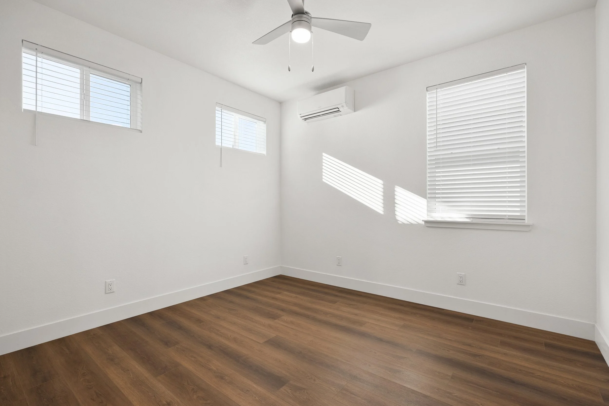 Empty room with white walls, three small windows with blinds, a ceiling fan, wooden floor, and an air conditioner.