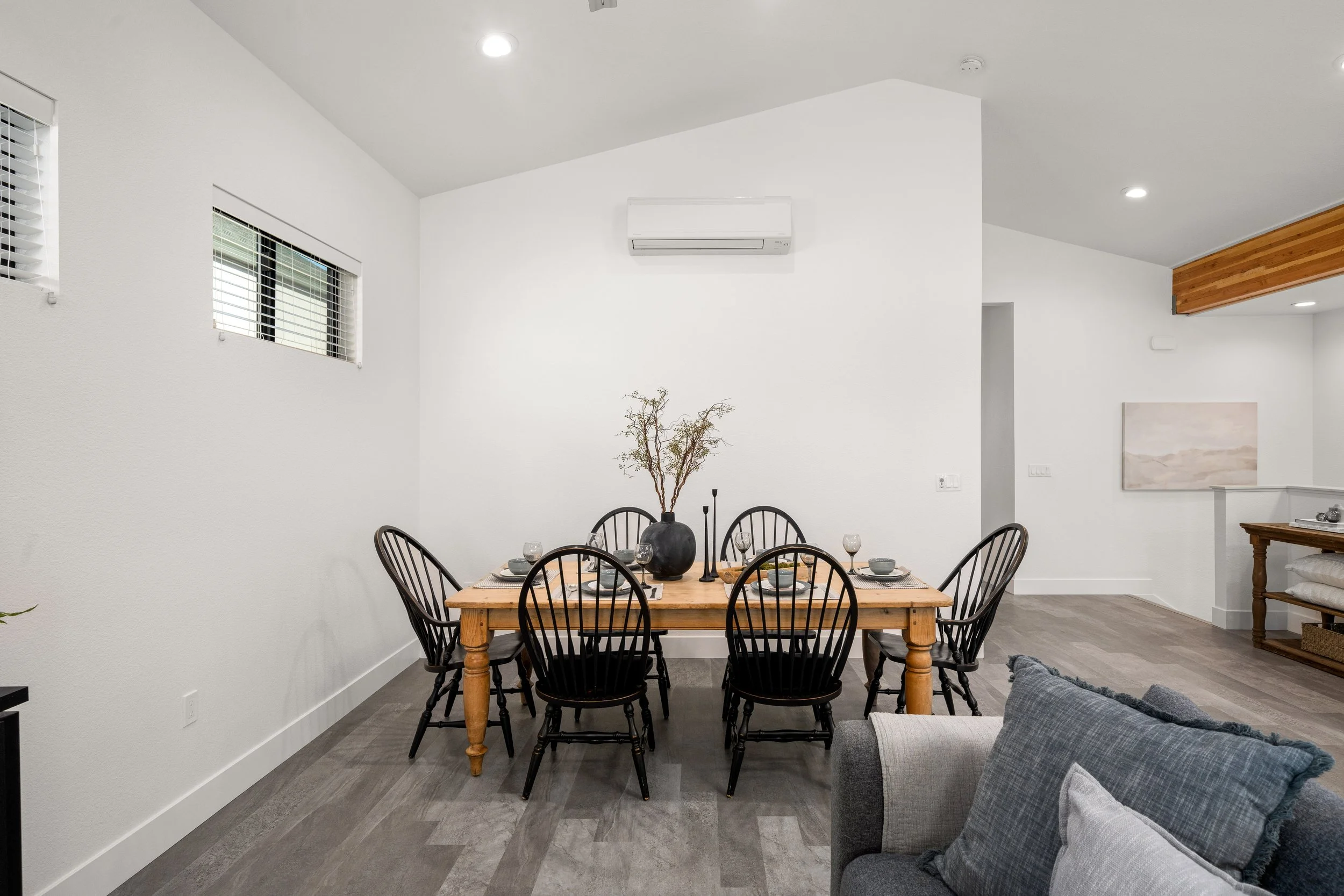 Dining room with a wooden table set for six, black chairs, a black vase with dried branches, white walls, and windows with white blinds.