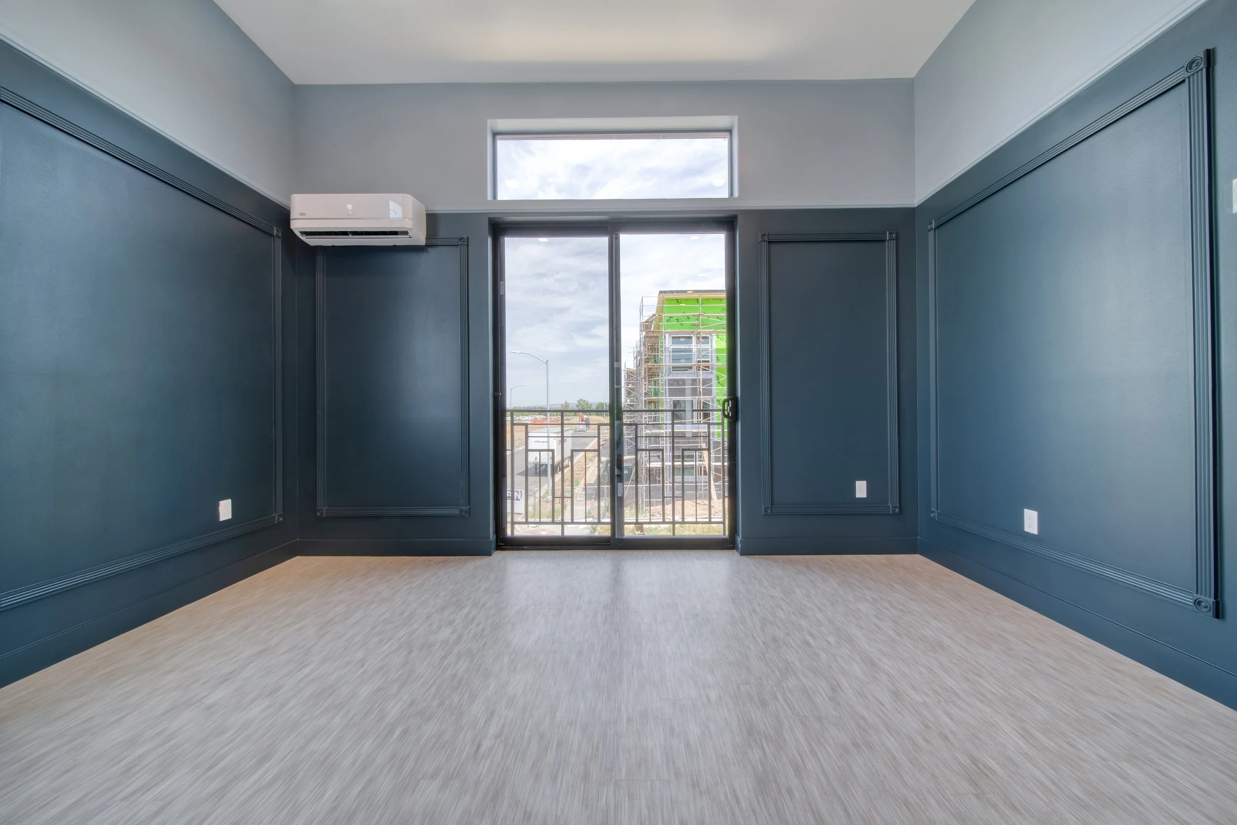Empty room with blue walls, a sliding glass door leading to a balcony, and a small window above the door, with light-colored hardwood flooring.