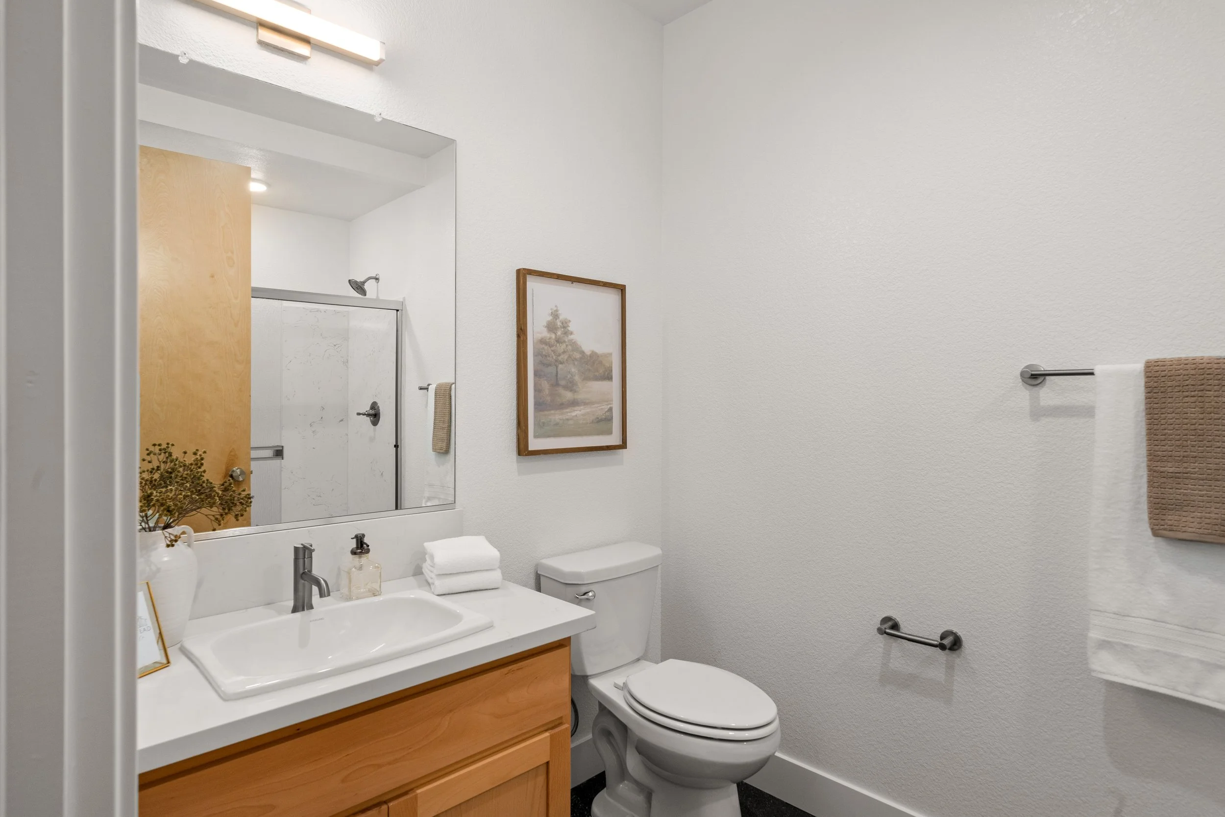 A small bathroom with a wooden vanity, a white sink, a mirror, a toilet, a framed landscape picture on the wall, and a towel rack with a brown towel.
