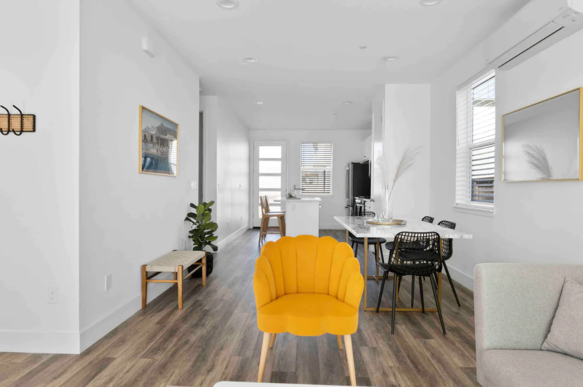 Living room with white walls, hardwood floors, yellow armchair, gray sofa, dining table with black chairs, TV, windows, and modern decor.