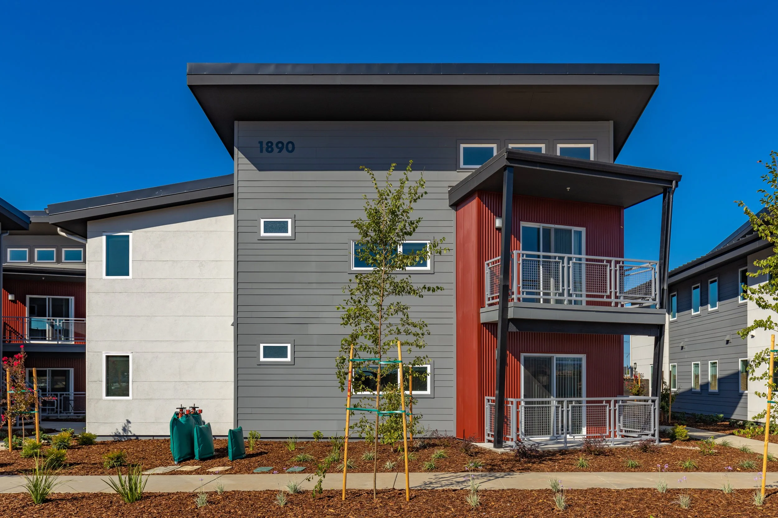 Modern multi-family residential building with gray, white, and red exterior siding, balconies, small landscaped yard, and a clear blue sky.