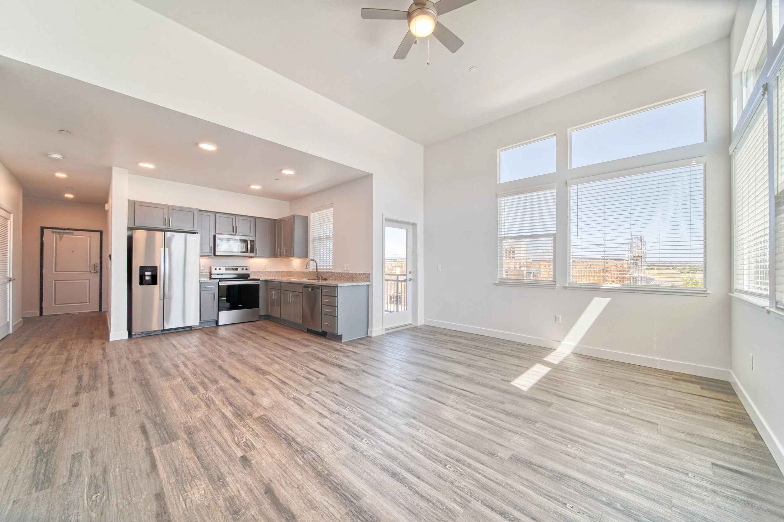 Empty living space with large windows, wood flooring, kitchen area with gray cabinets, stainless steel appliances, and a ceiling fan.