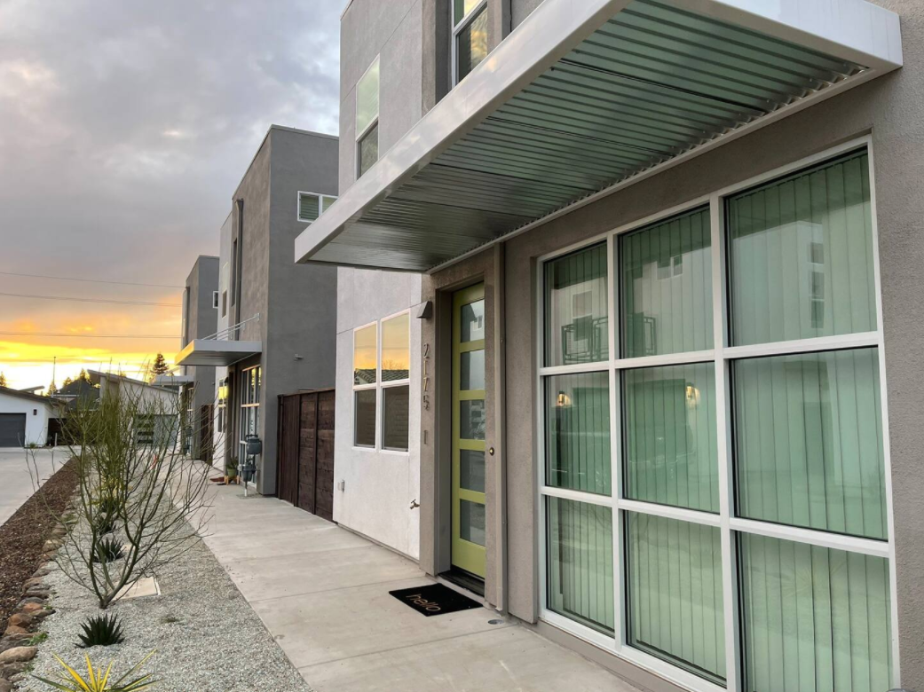 Modern townhouse with a green front door, large window with vertical blinds, and a small porch with overhang, during sunset.
