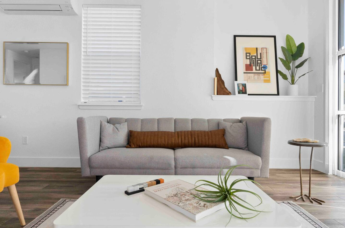 Modern living room with a light gray sofa, a brown pillow, a white coffee table, a side table, and wall art, with white walls and a window with blinds.