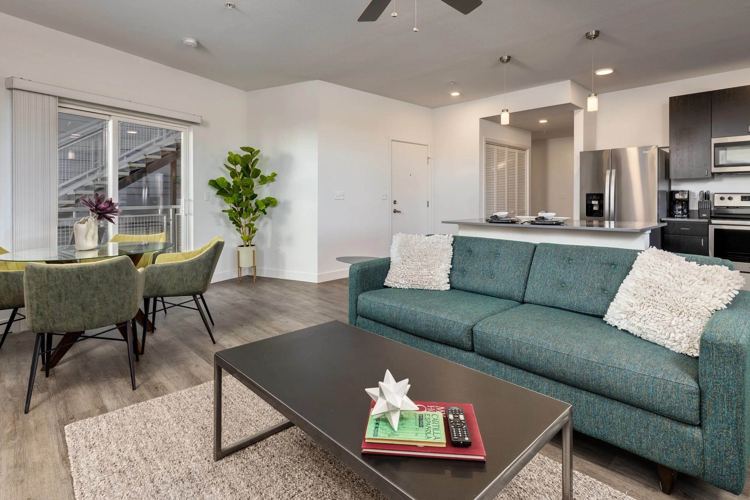 Open-concept living room and kitchen with teal sofa, dining area with green chairs, potted plant, and stainless steel appliances.
