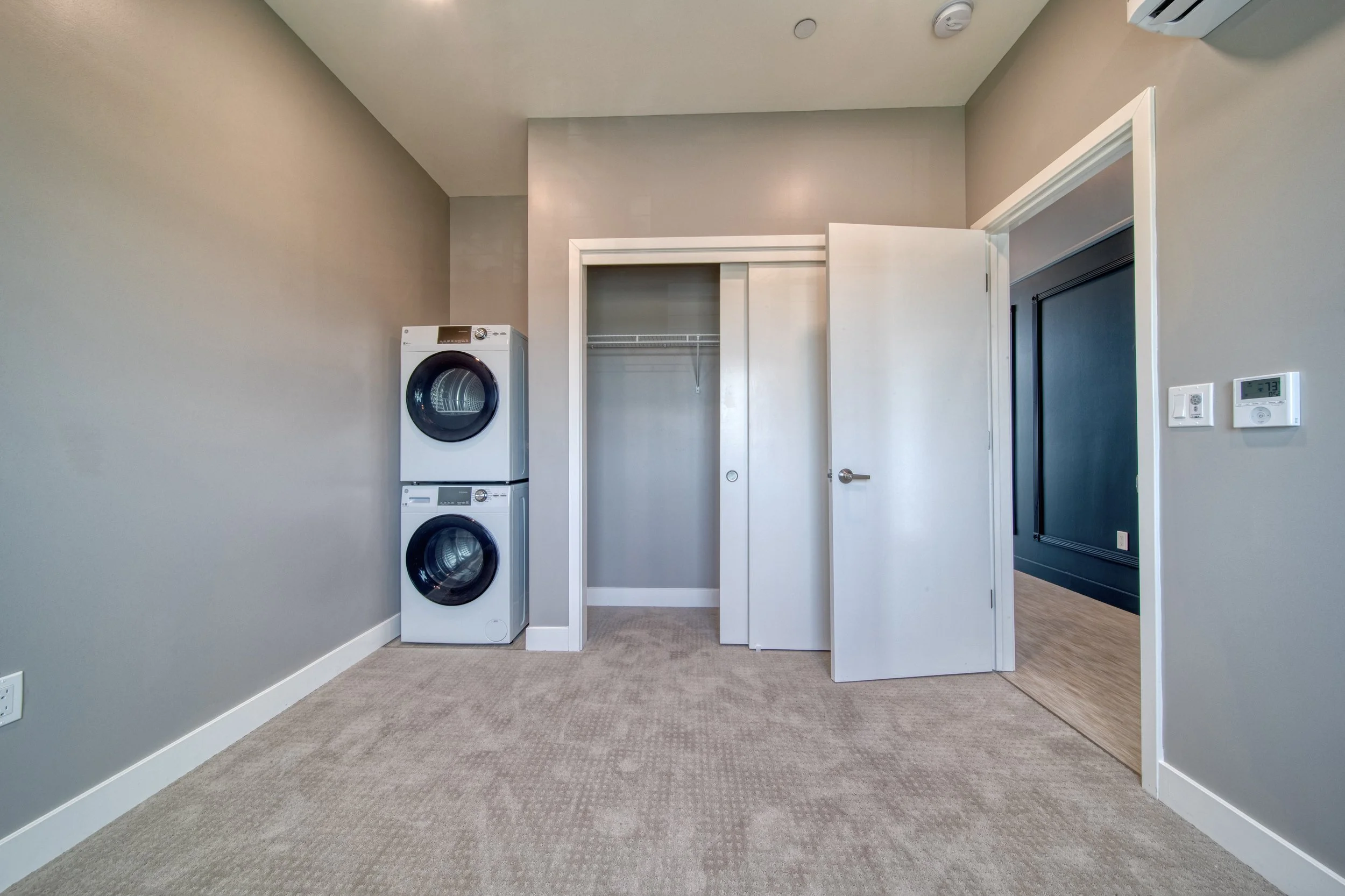 Empty laundry room with stacked washer and dryer, open closet door with hanging rod, and light-colored carpeted floor.
