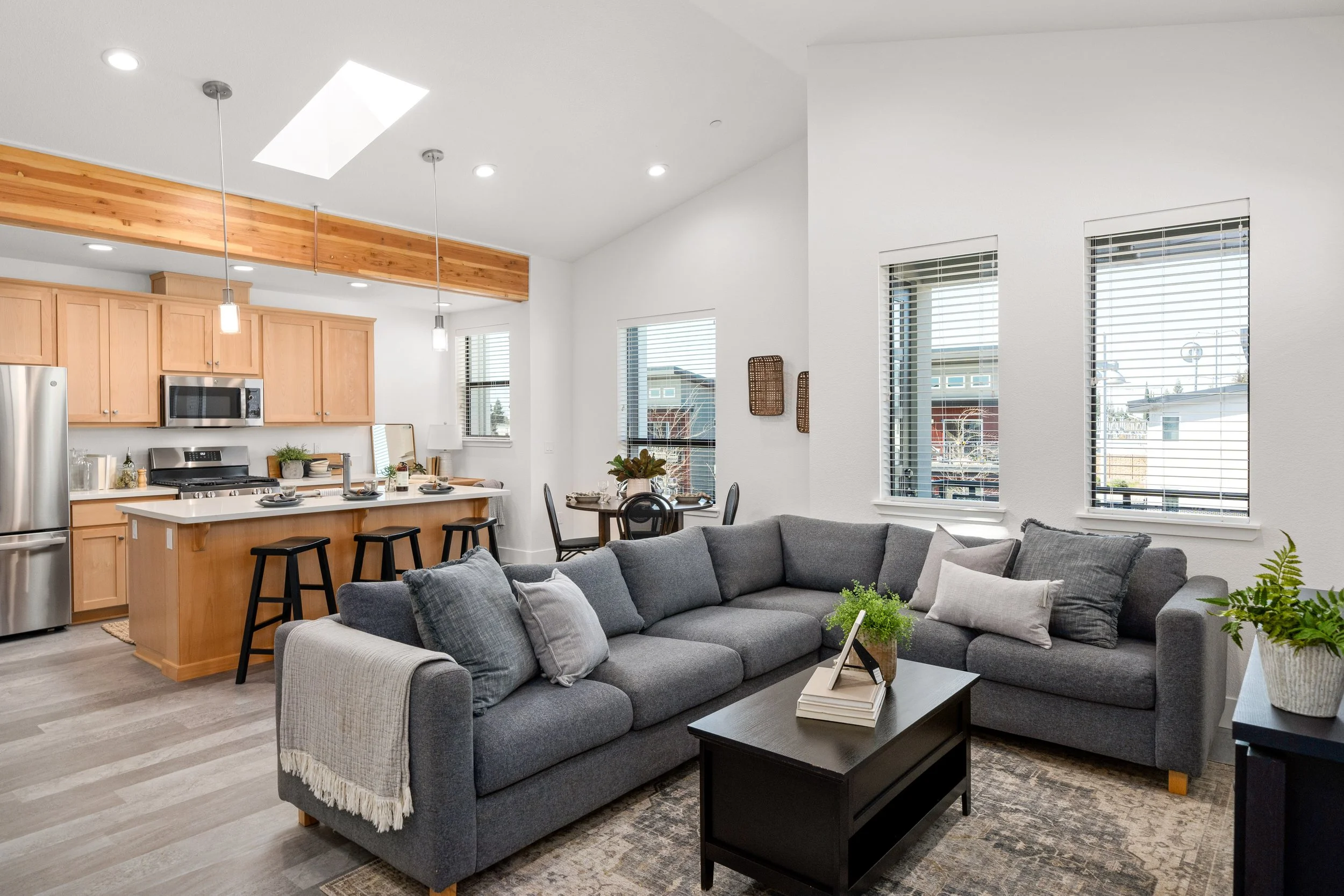 Open-concept living room and kitchen with light wood cabinets, stainless steel appliances, and a large gray sectional sofa with throw pillows. Multiple windows with blinds, potted plants, and a wooden dining table with four chairs are visible.