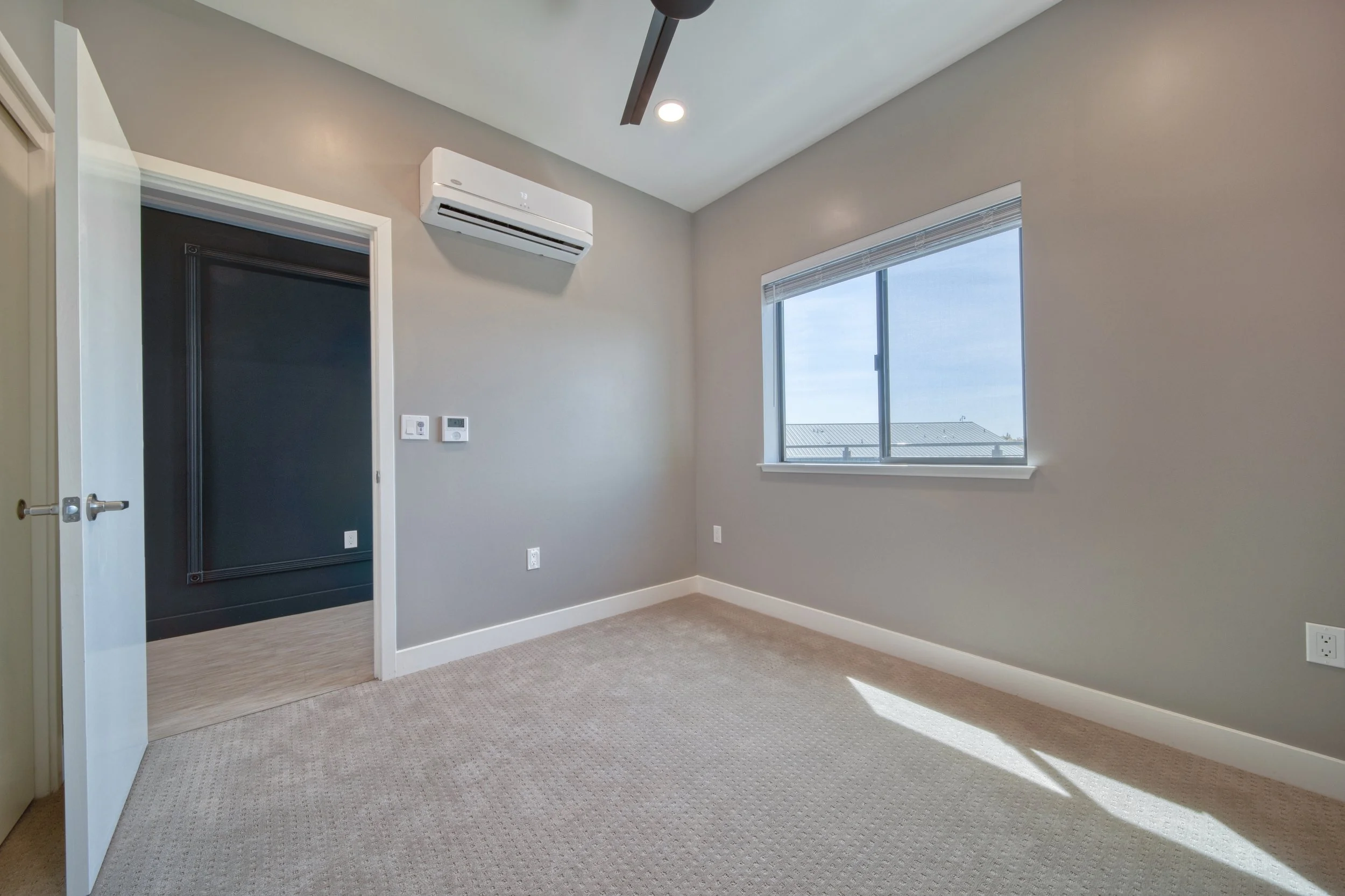 Empty room with gray walls, beige carpet, a window, wall-mounted air conditioner, and a partially open door leading to an adjacent darker room.