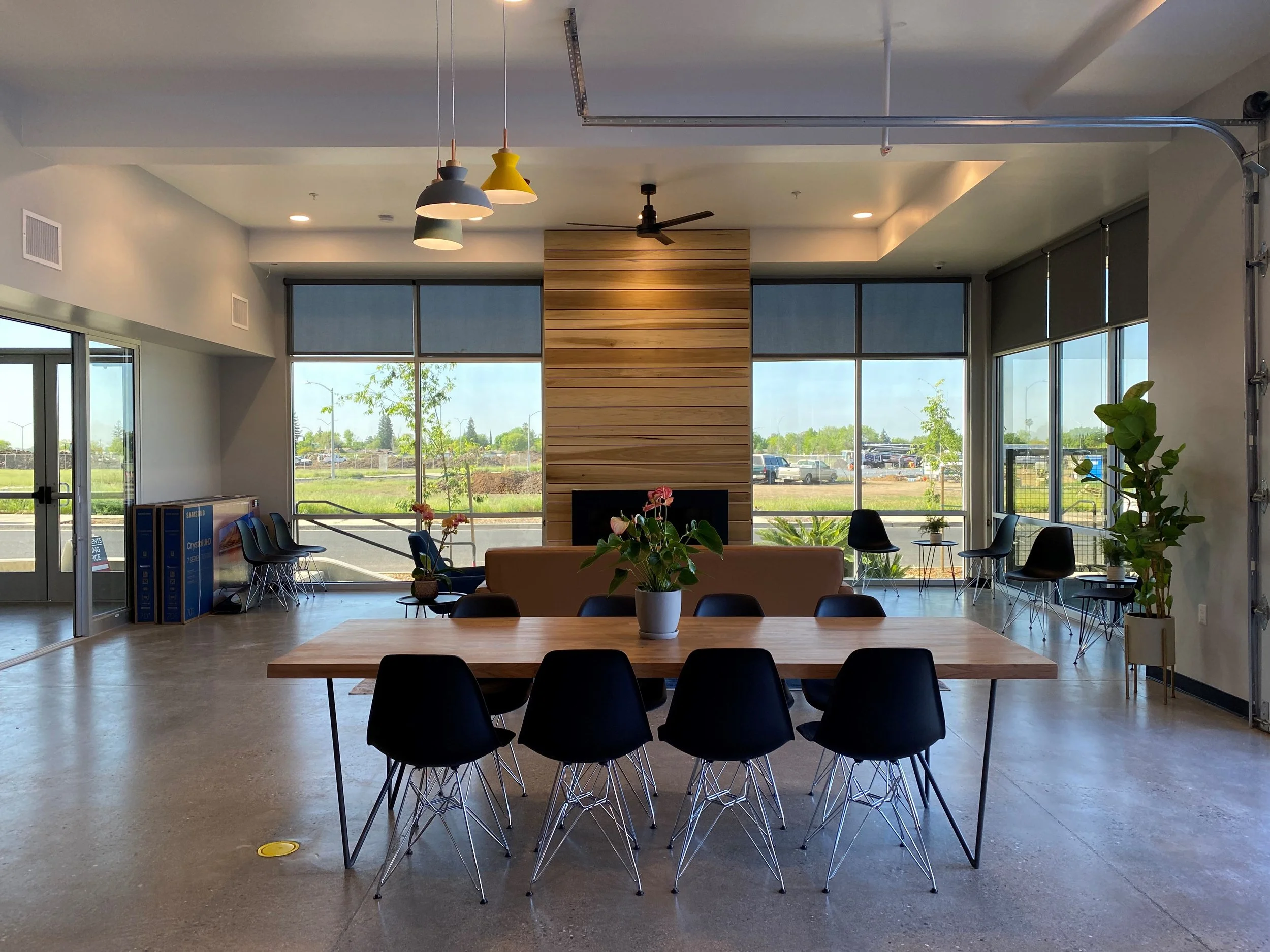 Bright modern lobby with large windows, a wooden accent wall with a fireplace, a central table with chairs, hanging pendant lights, and potted plants.