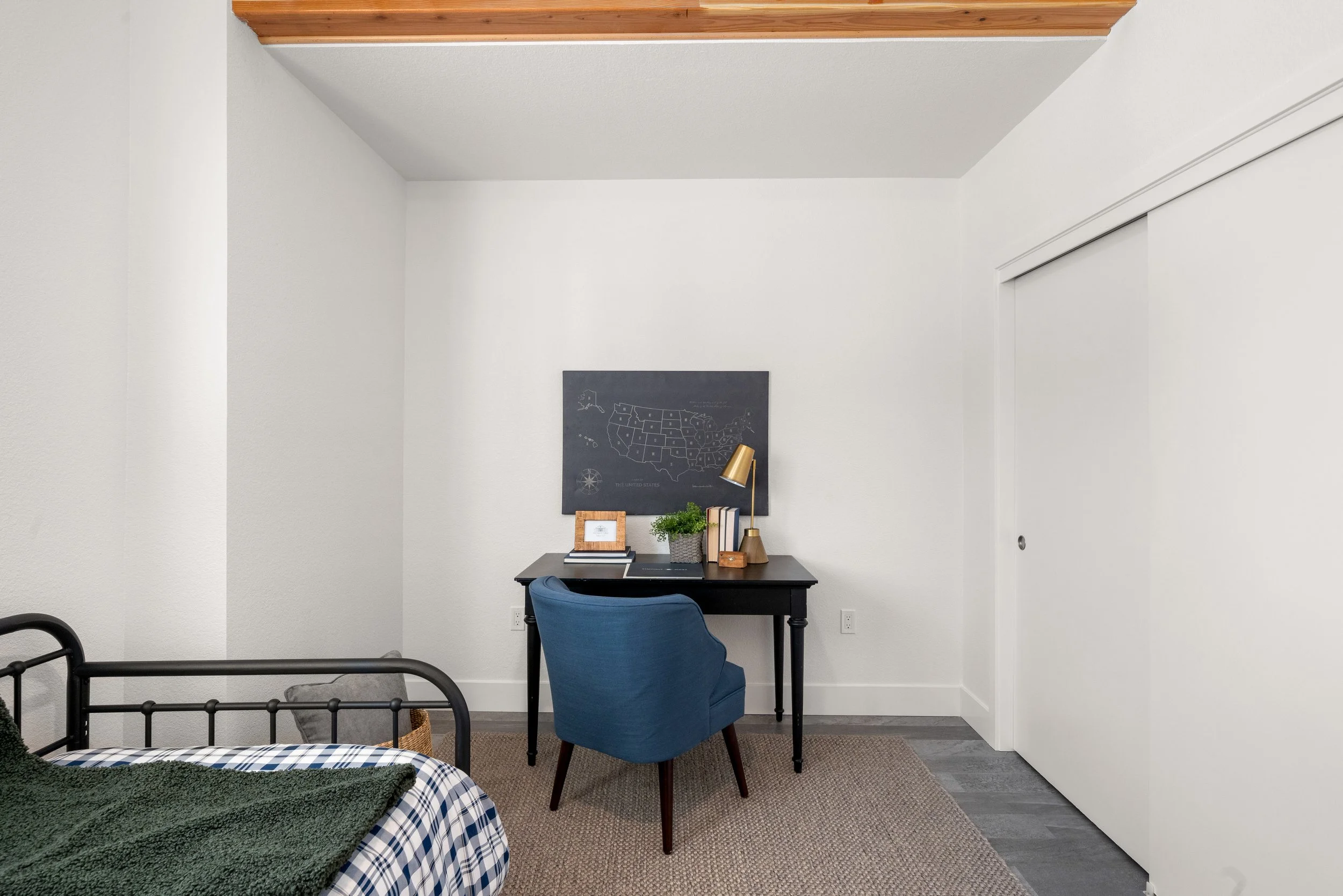 A small bedroom with a black desk and a blue upholstered chair, a black chalkboard with a map of the United States, a potted plant, lamp, clock, and books on the desk, a bed with a black frame and checkered sheets, a green blanket, a basket near the 