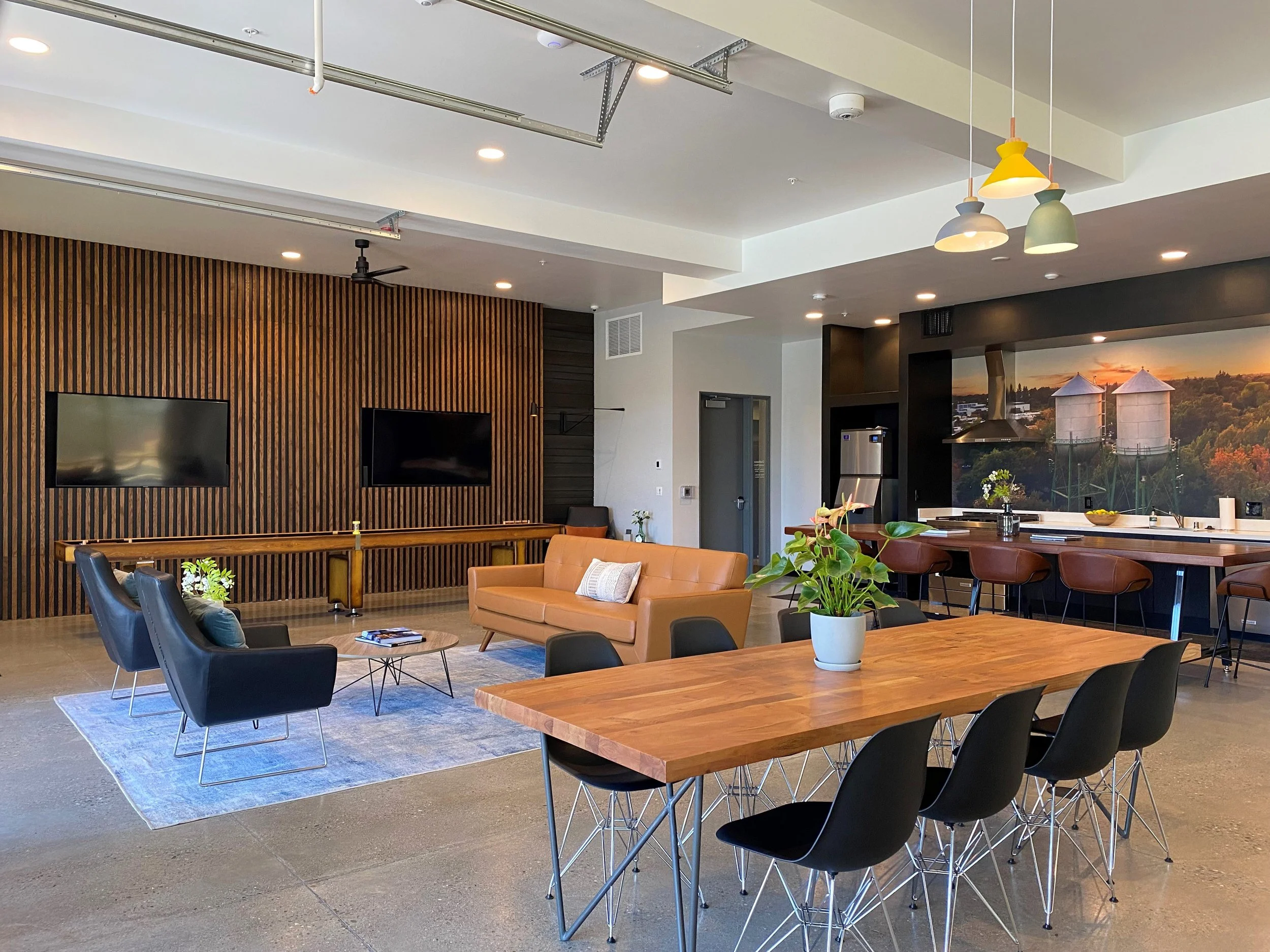 Modern living room with a wooden accent wall, two flat-screen TVs, leather and fabric chairs, a rug, a coffee table, a wooden dining table with black chairs, a potted plant, and a bar area with barstools and a large mural of a farm scene with silos a