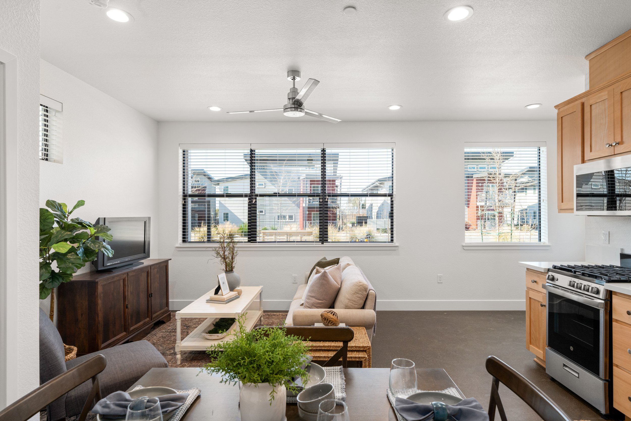 Bright living area with white walls, three large windows with blinds, a ceiling fan, and a kitchen with wooden cabinets and a stainless steel stove. There is a sofa, a TV on a wooden stand, and a dining table with a plant centerpiece and place settin