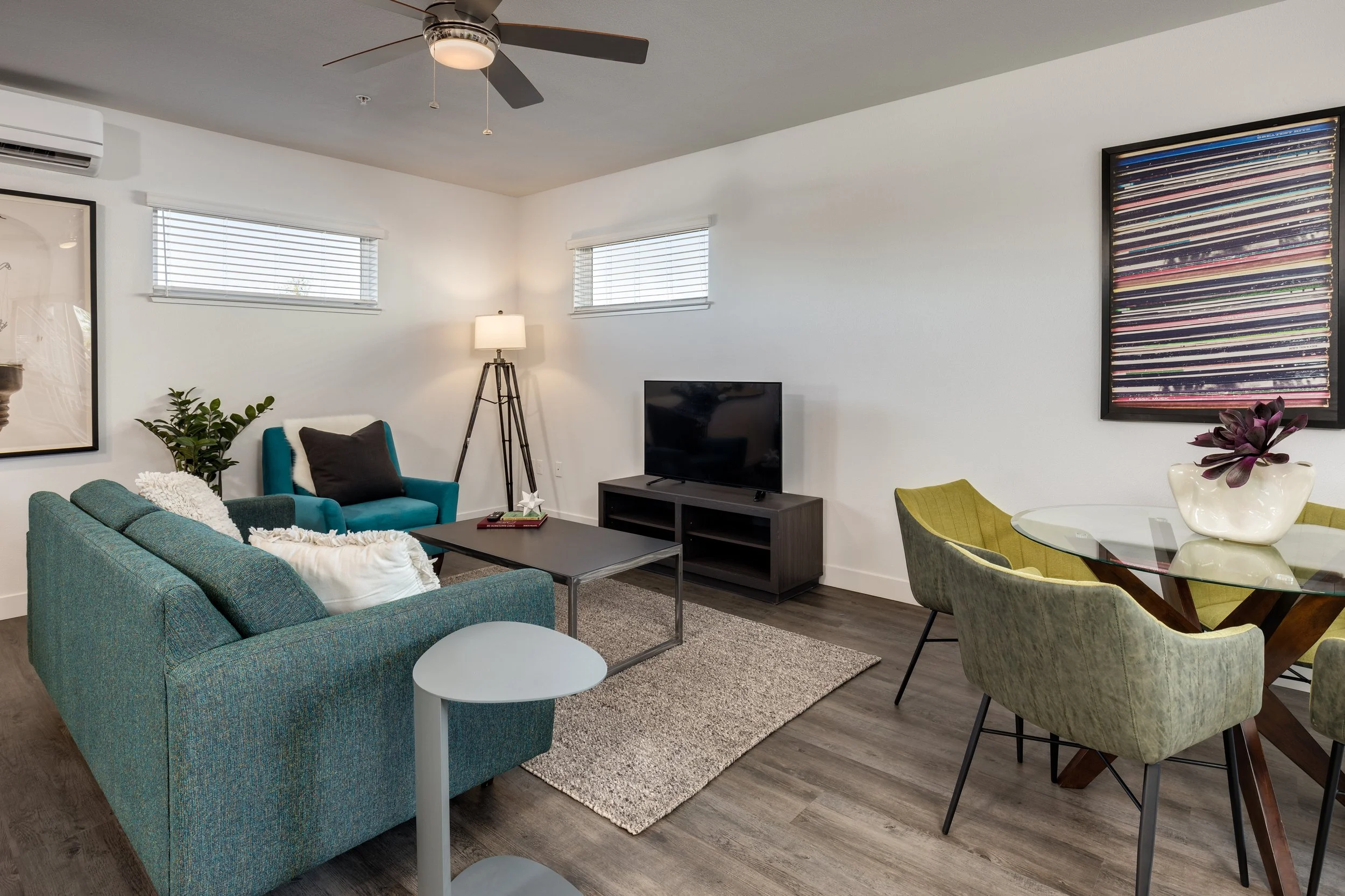 Modern living room with gray sofa, teal armchair, black coffee table, TV on dark wood stand, green dining chairs, glass-top dining table, abstract art on wall, potted plant, ceiling fan, window blinds, and wooden flooring.