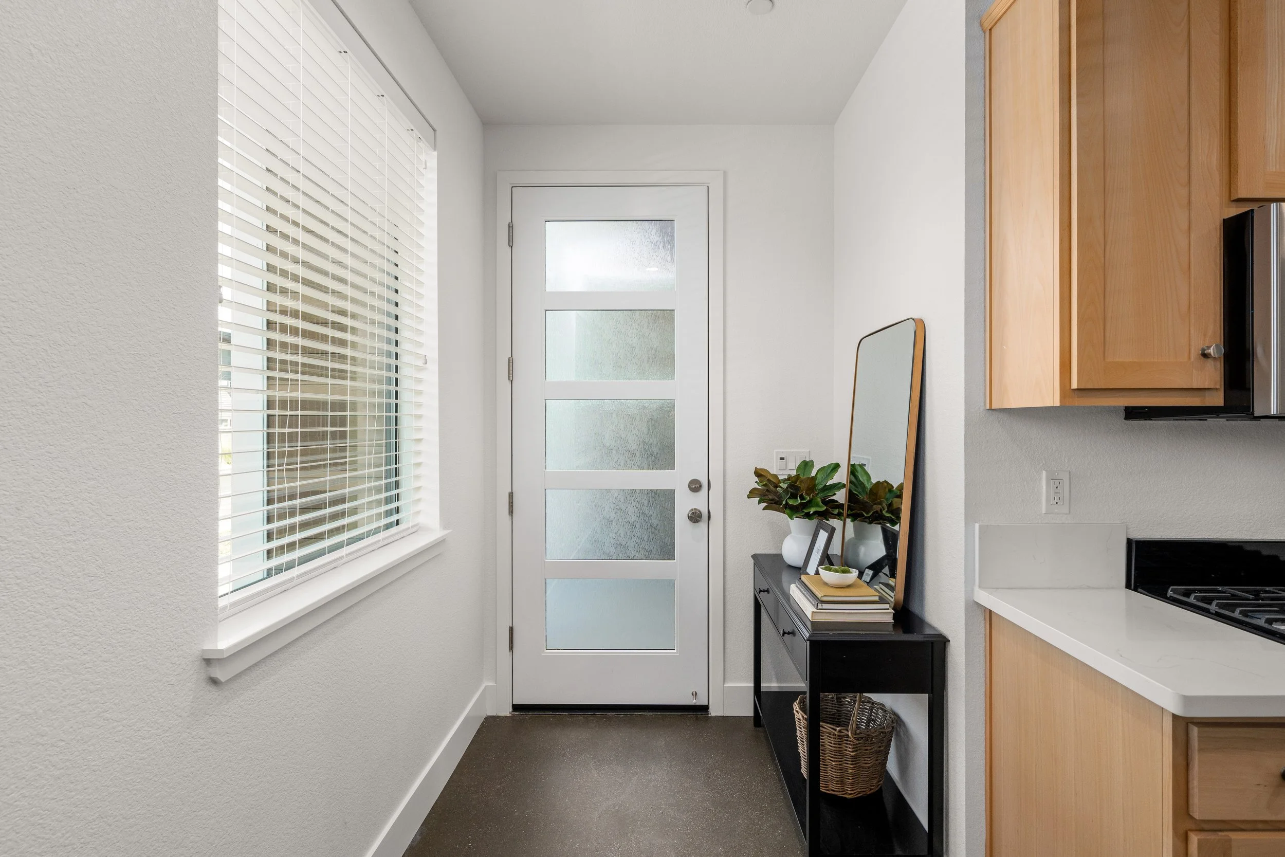 Interior view of a kitchen entryway with a white door, frosted glass panels, a window with blinds, a black slim table with decorative items, a large mirror, a houseplant, and kitchen cabinets.