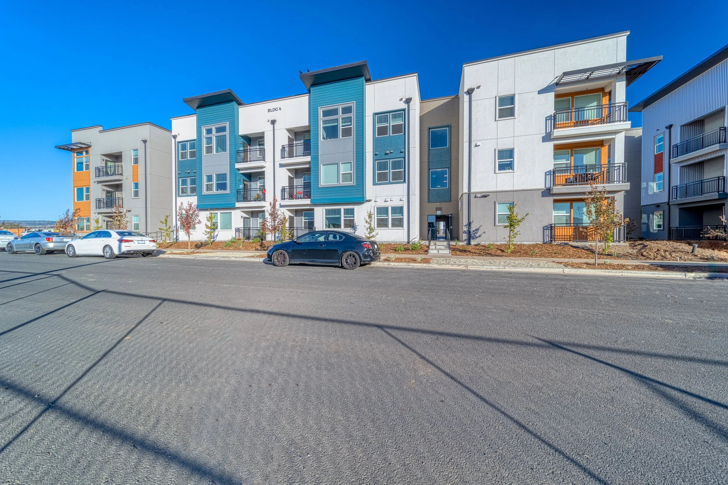 Exterior view of a modern, multi-story apartment building with a parking lot in front, under a clear blue sky.