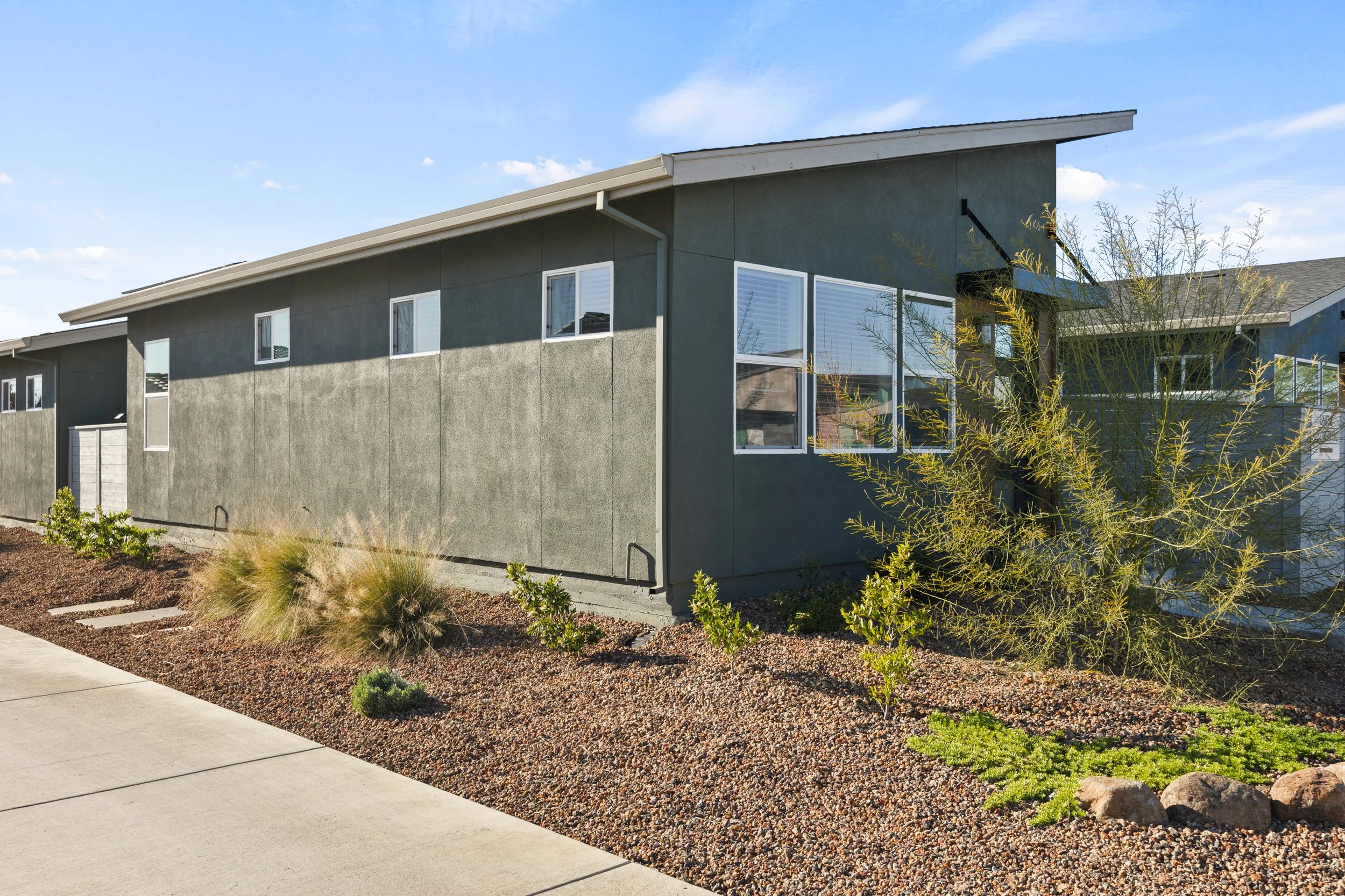 Modern two-story residential building with dark gray exterior walls, large windows, a sloped roof, and desert landscaping with shrubs and rocks in the foreground under a blue sky.