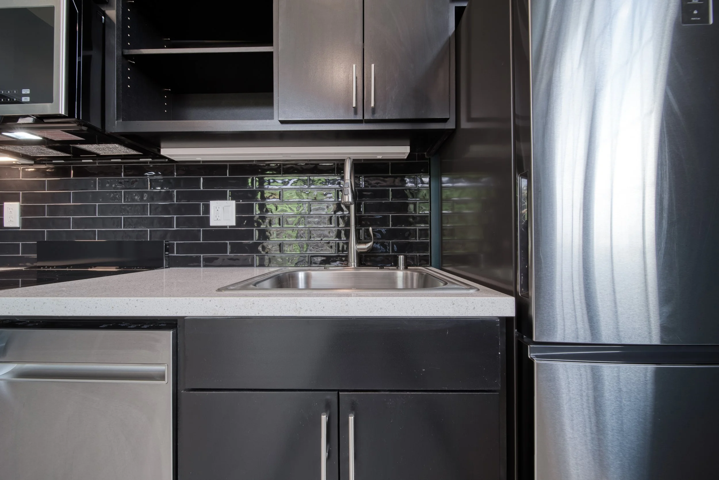 Modern kitchen with black cabinets, stainless steel refrigerator, black backsplash, and white countertop with a sink.