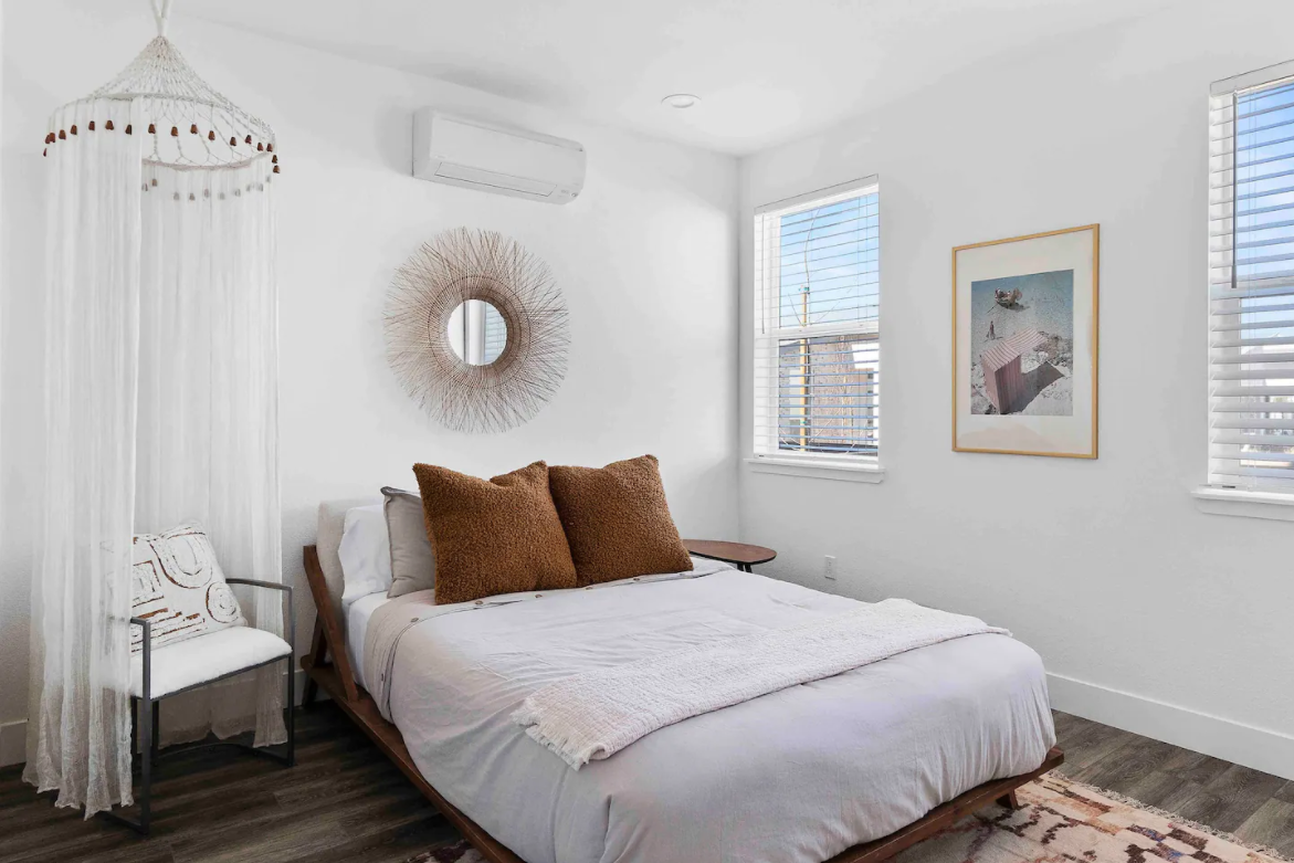 A bedroom with white walls, a bed with white bedding and brown pillows, two windows with blinds, framed art on the wall, a sitting chair, and a circular mirror decorated with a sunburst design.