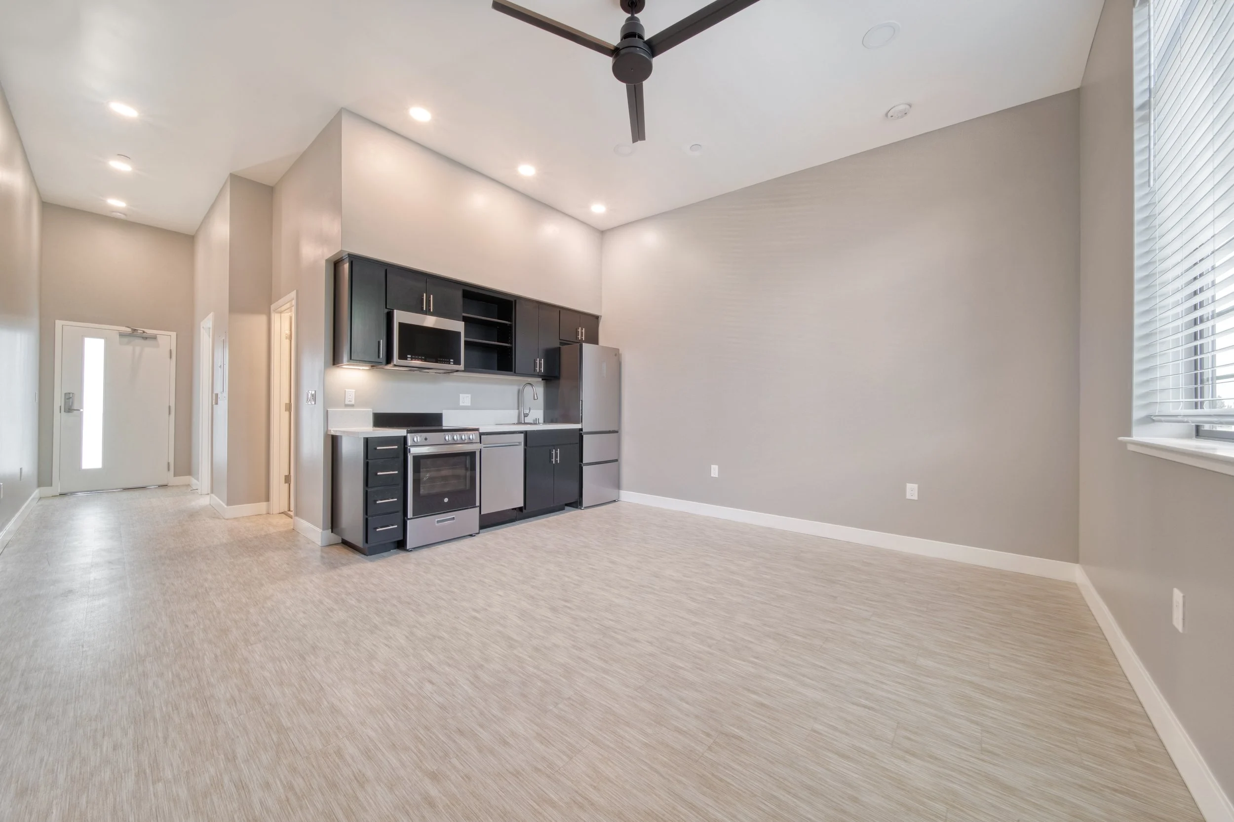 Empty modern kitchen and dining area with gray walls, wooden flooring, black cabinets, stainless steel appliances, and a ceiling fan.