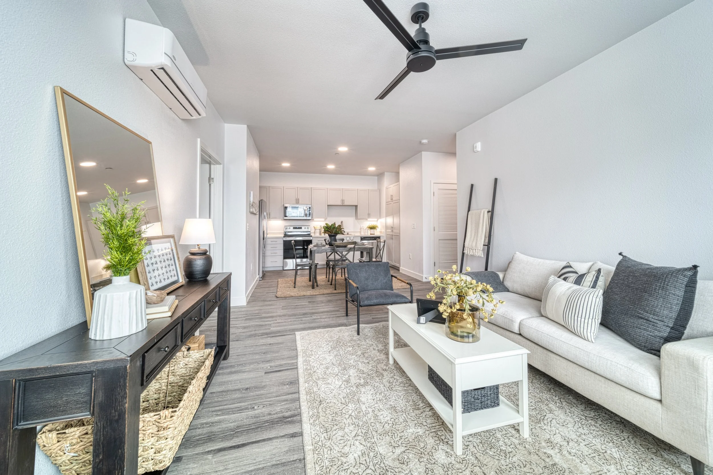 Open-concept living room with white sofa, black armchair, white coffee table, wall decor, and kitchen in the background