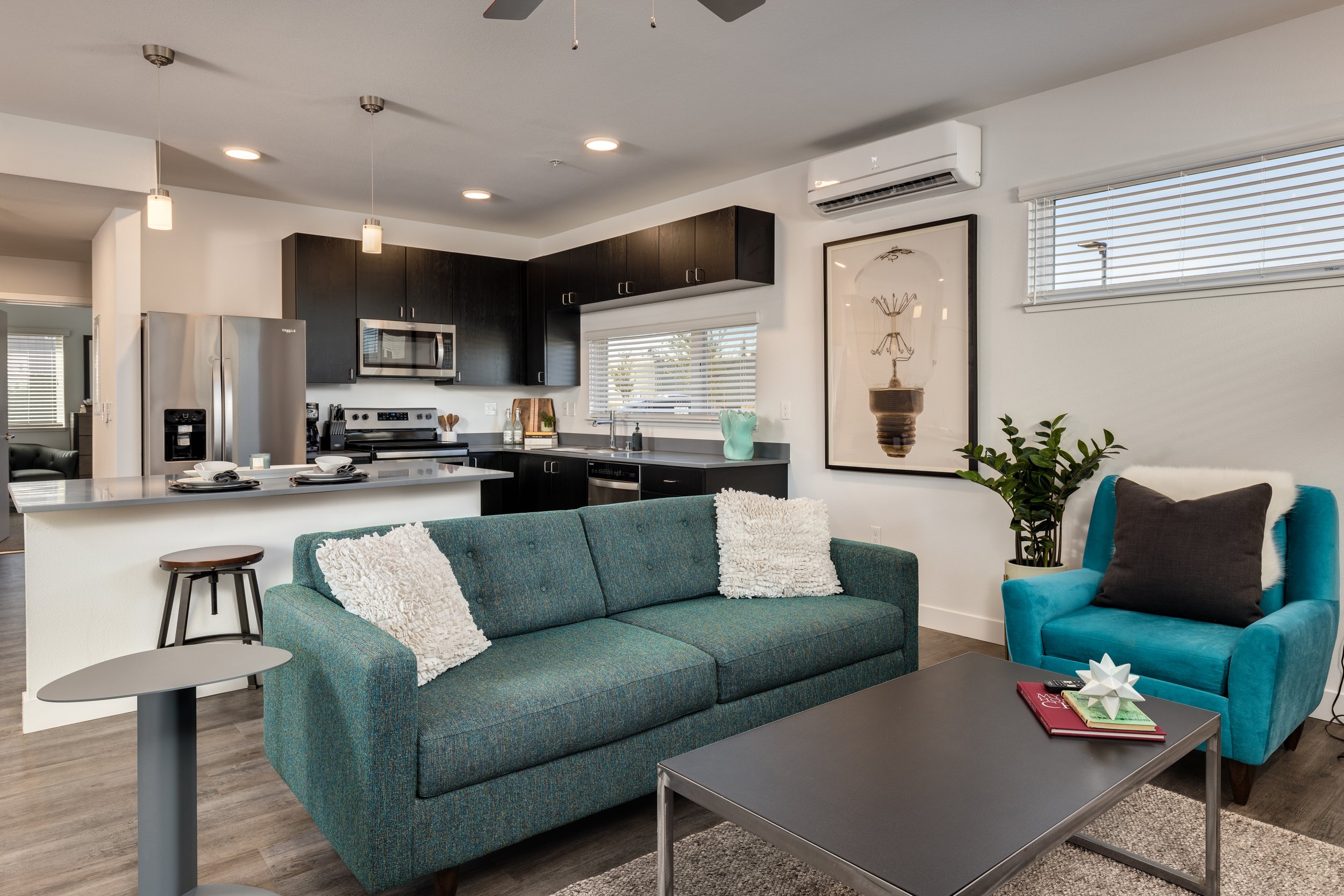 Living room with teal sofa and armchair, coffee table with books and decorative object, potted plant, and artwork on the wall. Open kitchen with dark cabinets, stainless steel appliances, and pendant lighting.
