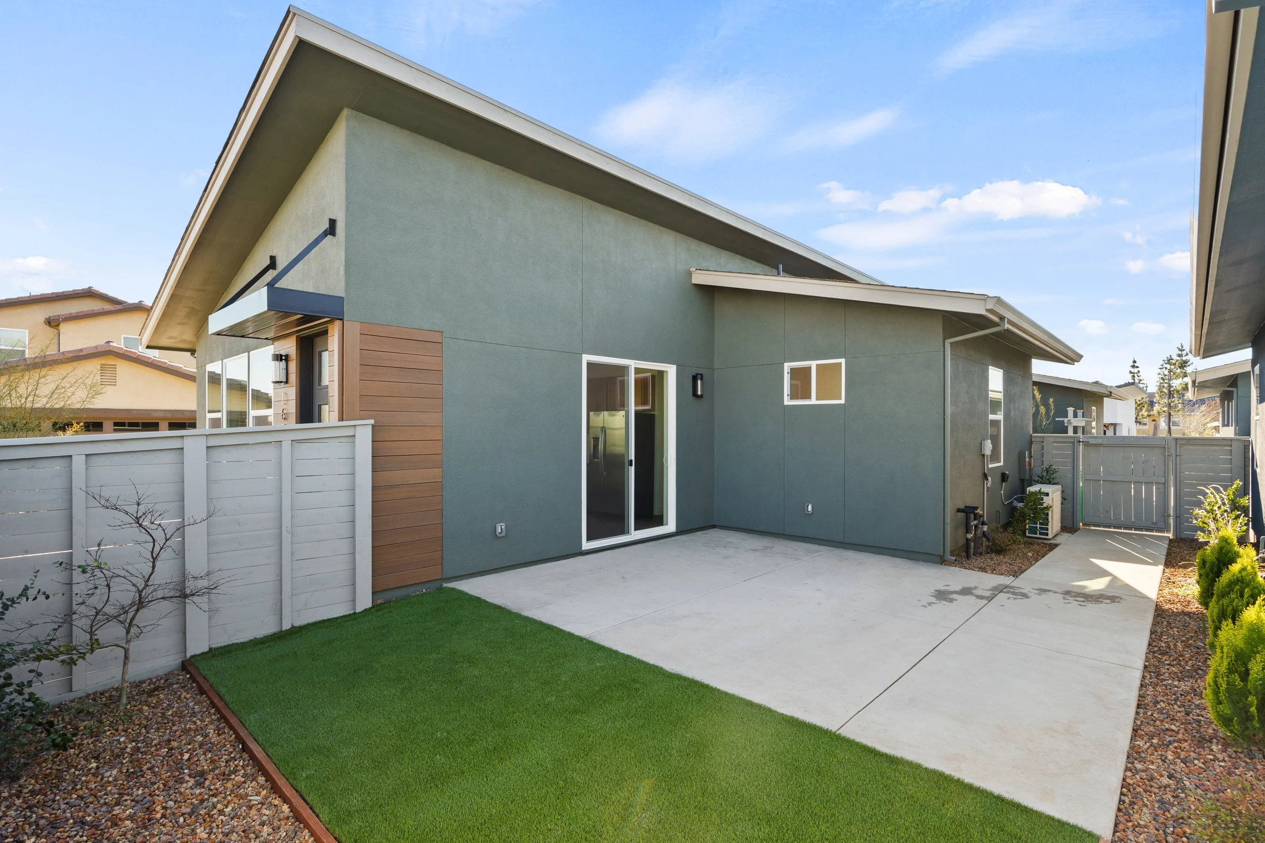 Modern house backyard with concrete patio, small patch of artificial grass, wooden fence, and overcast sky.