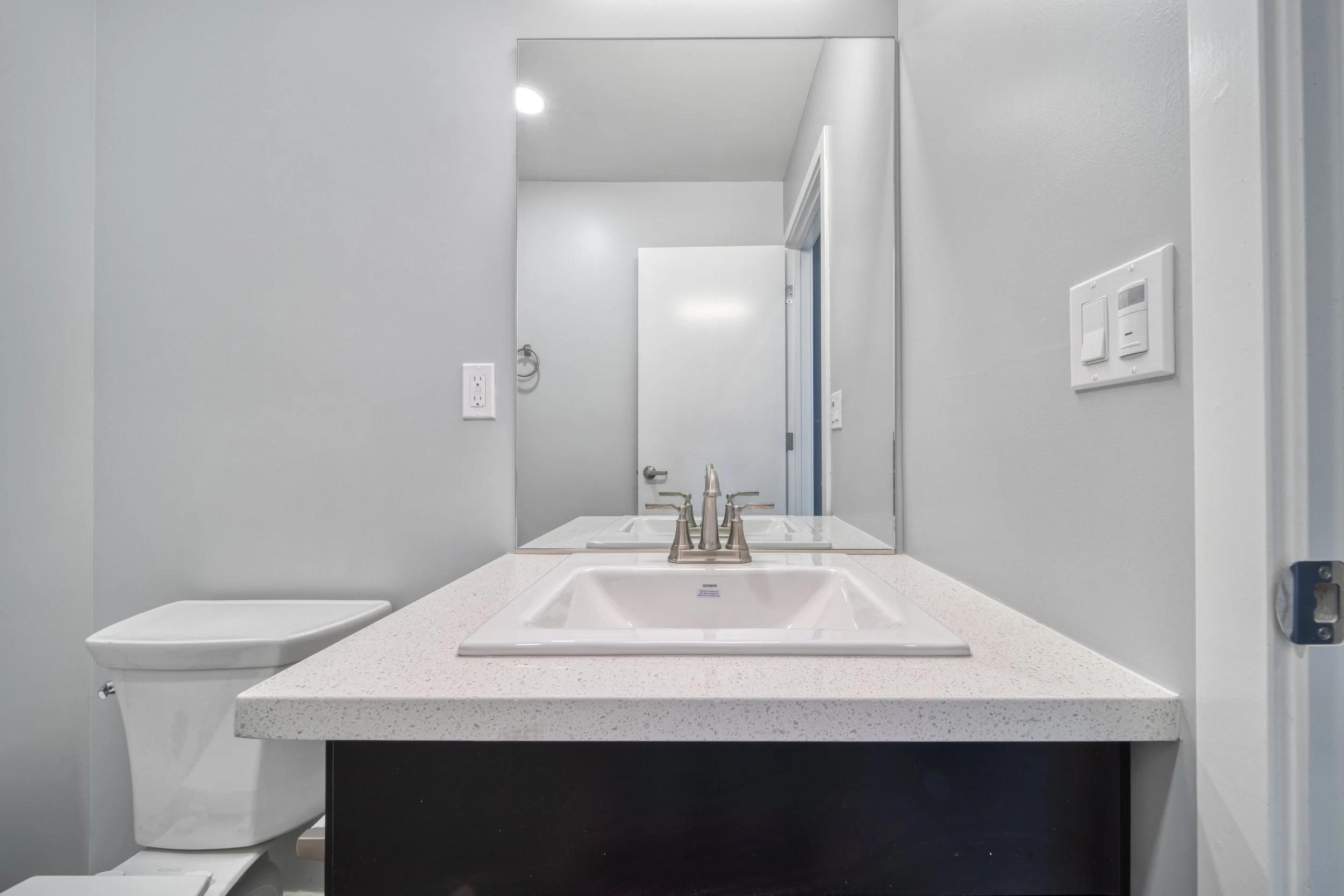 Bathroom with a single sink, faucet, large mirror, and a toilet in the corner. The walls are painted light gray.