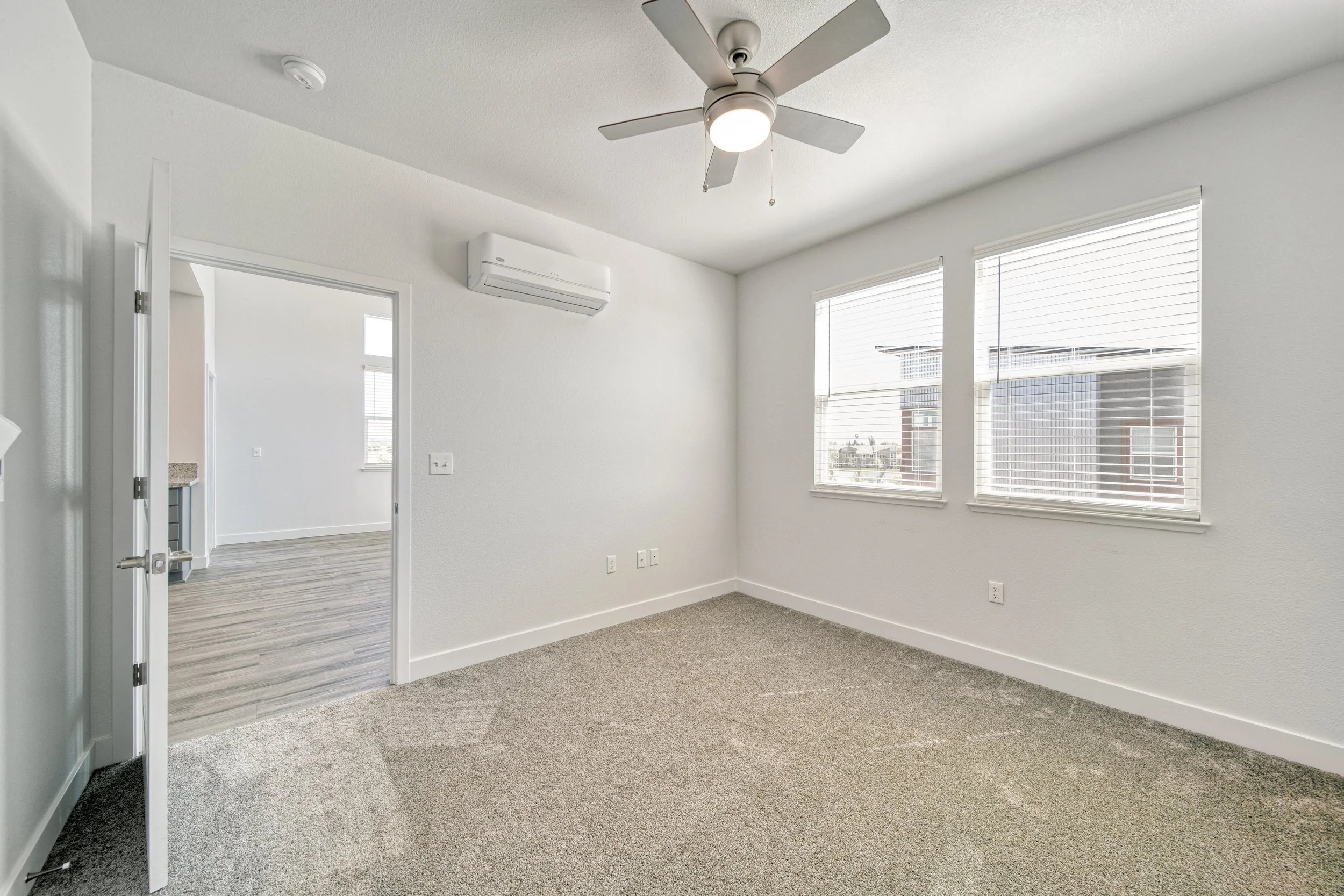 Empty room with white walls, beige carpet, two windows with white blinds, a ceiling fan with a light, and an air conditioning unit mounted on the wall.