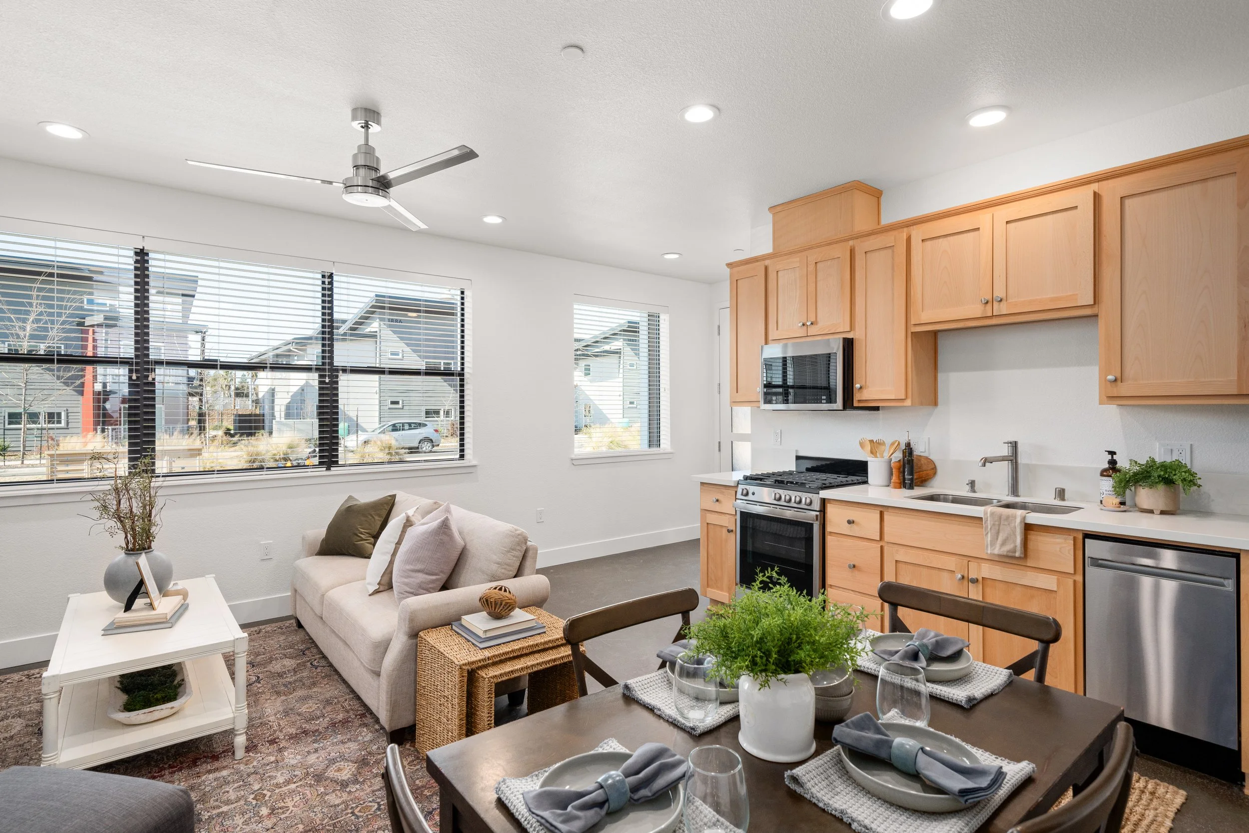 Open-concept living room and kitchen with large windows, beige sofa with pillows, dining table with place settings, and light wood kitchen cabinets with stainless steel appliances and green plants.