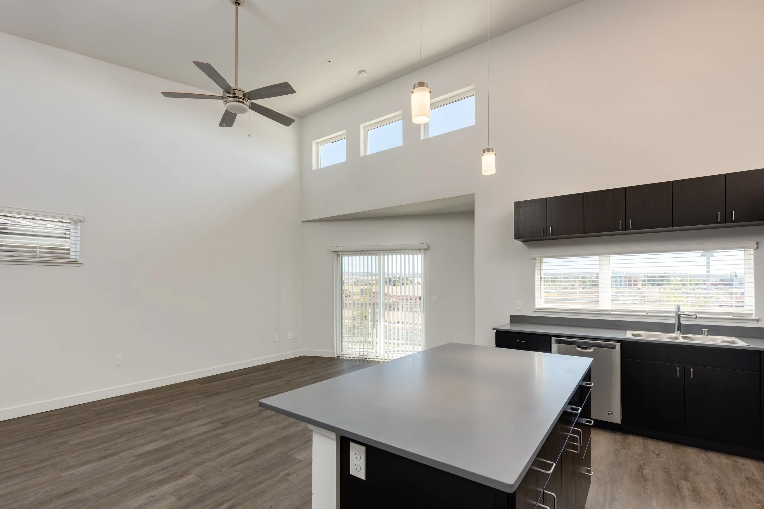 An open spacious kitchen and living area with high ceilings, a ceiling fan, pendant lighting, dark cabinetry, a large window above the sink, and a glass sliding door leading outside.