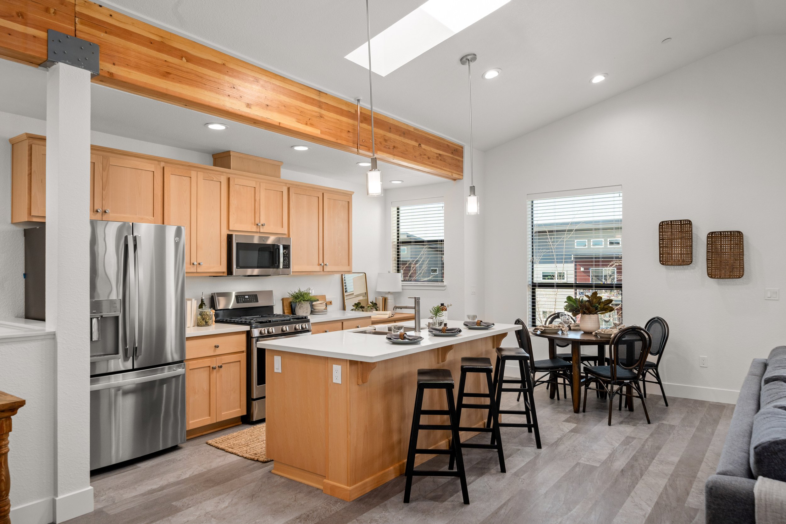 Open-concept kitchen and dining area with light wood cabinetry, stainless steel appliances, a white kitchen island with seating, and a dining table with four chairs. Spacious room with large windows and minimal decor.
