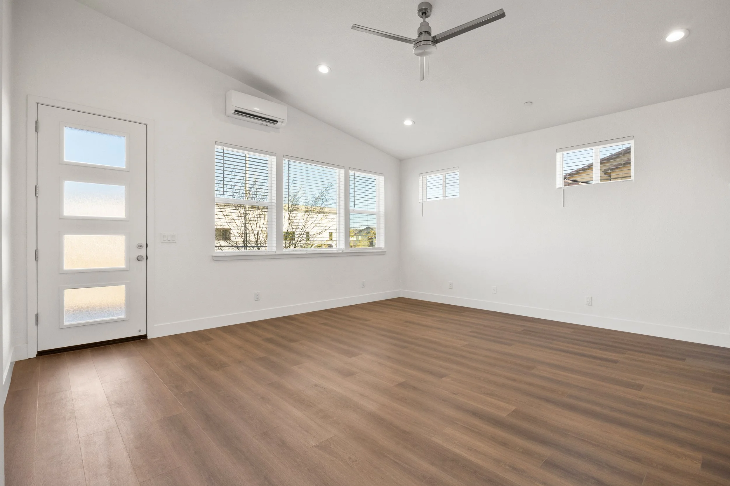Empty living room with white walls, wooden flooring, large windows with blinds, a ceiling fan, and recessed lighting.